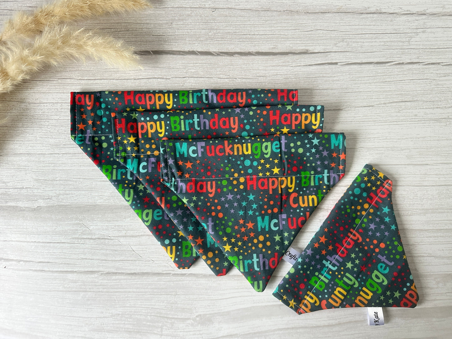Three handcrafted cotton pet bandanas by Crafts by Kate feature festive, humorous text on a starry dark background. Arranged fan-like on a light wood surface with dried grass, these "Happy Birthday C Mcnugget" bandanas are perfect for memorable moments.