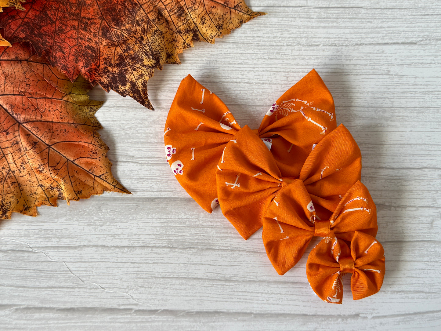 Three "Cotton Pet Bow with collar attachment - Dancing Skeletons" by Crafts by Kate are displayed on a light wooden surface next to two red and orange autumn leaves. Each bow features white skeletons on orange cotton and an easy elastic collar fastening.