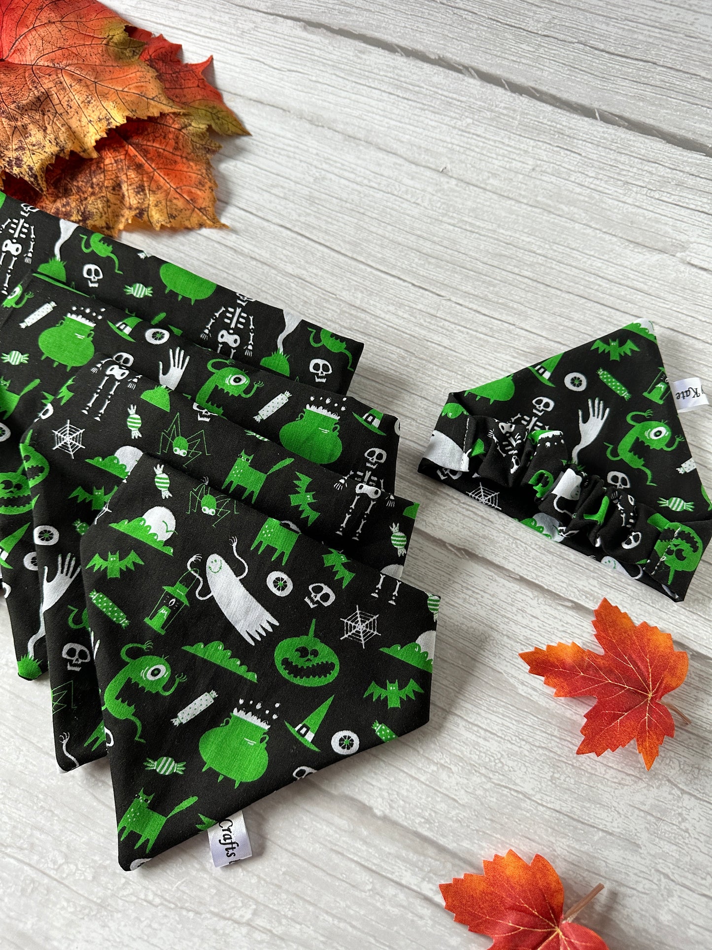 Crafts by Kate's Cotton Pet Scrunchie Bandana - Green Halloween features a handcrafted design with skeletons, pumpkins, bats, and monsters on cotton fabric. Displayed on a light wooden surface with autumn leaves.