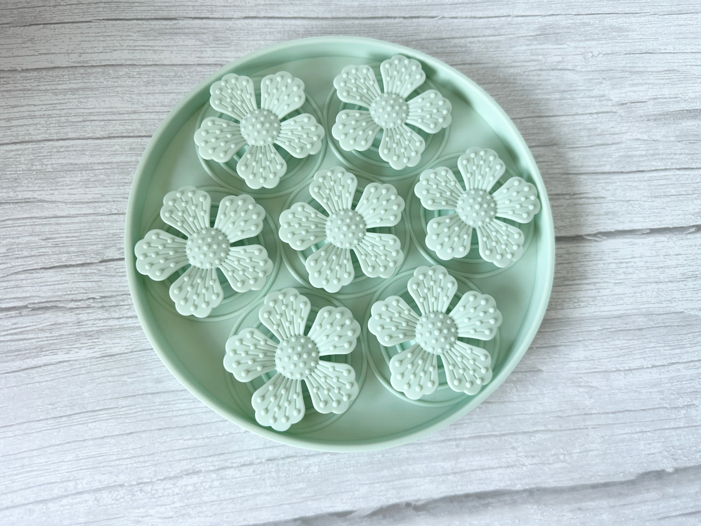 The Crafts by Kate Slow Feeder Pet Mat is a round, mint-colored tray featuring suction cups and securely holding six textured, mint flower shapes with detailed petals on a light wood background.