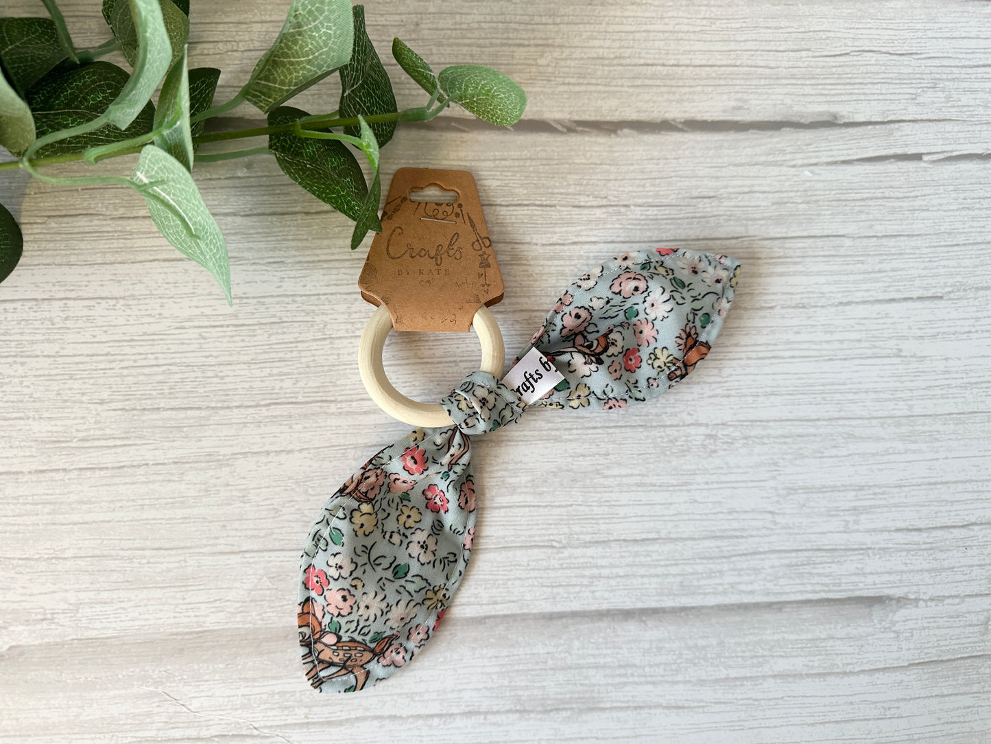 Part of the "Floral Bambi" baby gift set by Crafts by Kate, this wooden teething ring features ultra-soft floral-patterned cotton bunny ears and rests on a white wooden surface next to a small leafy plant.