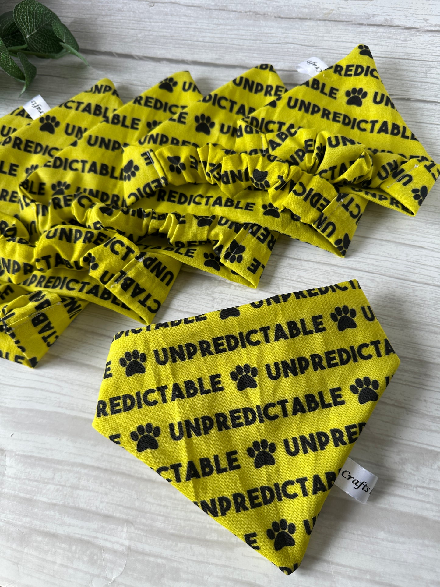 The Cotton Pet Scrunchie Bandana - Unpredictable by Crafts by Kate is a yellow handcrafted set with black paw prints and "UNPREDICTABLE" in bold, shown on a light wooden surface.