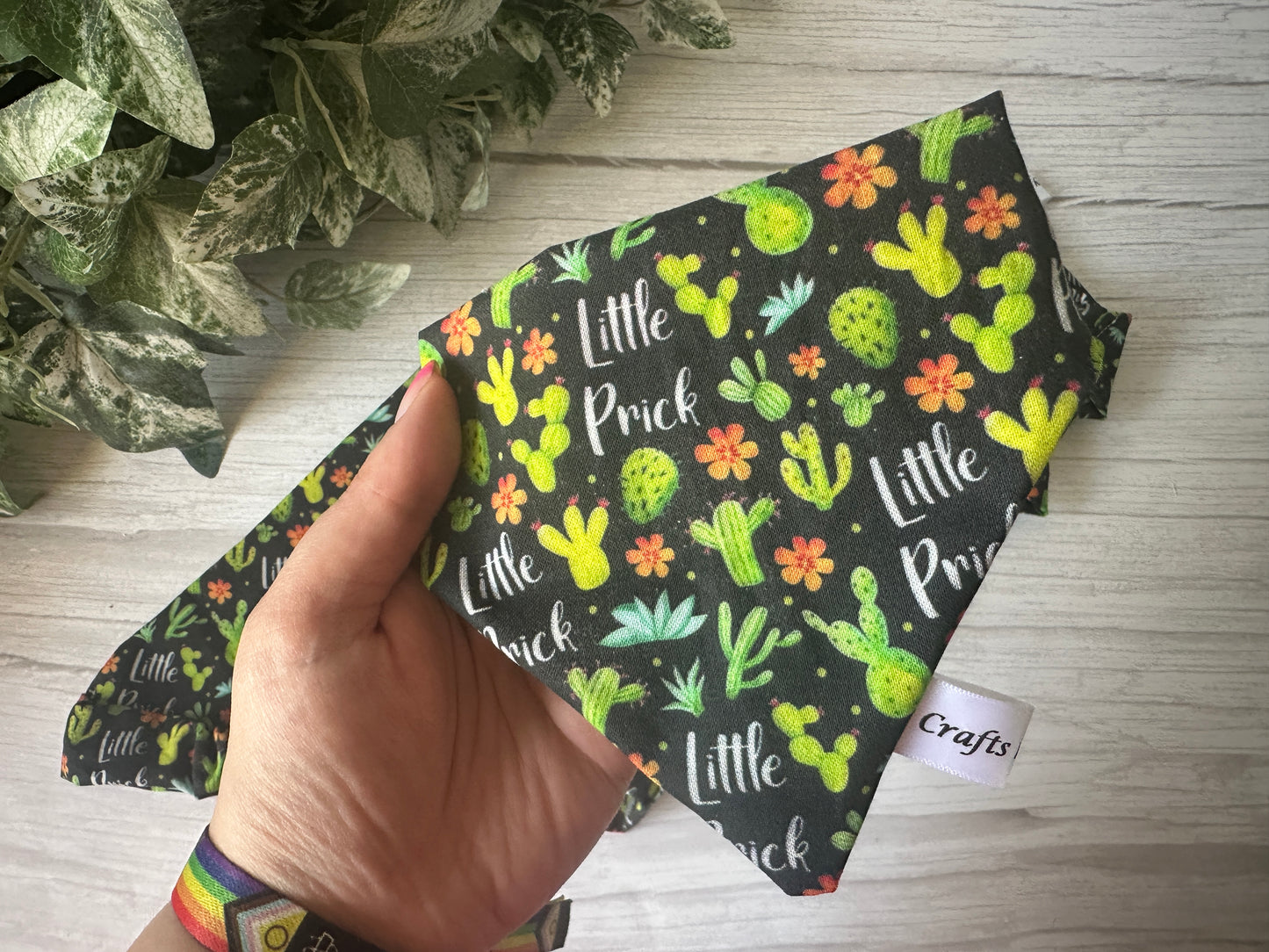 A hand holds the Cotton Pet Scrunchie Bandana - Little Prick by Crafts by Kate, featuring colorful cacti, flowers, and “Little Prick” lettering. A small white "Crafts" tag is sewn on. Green leaves are seen in the background.