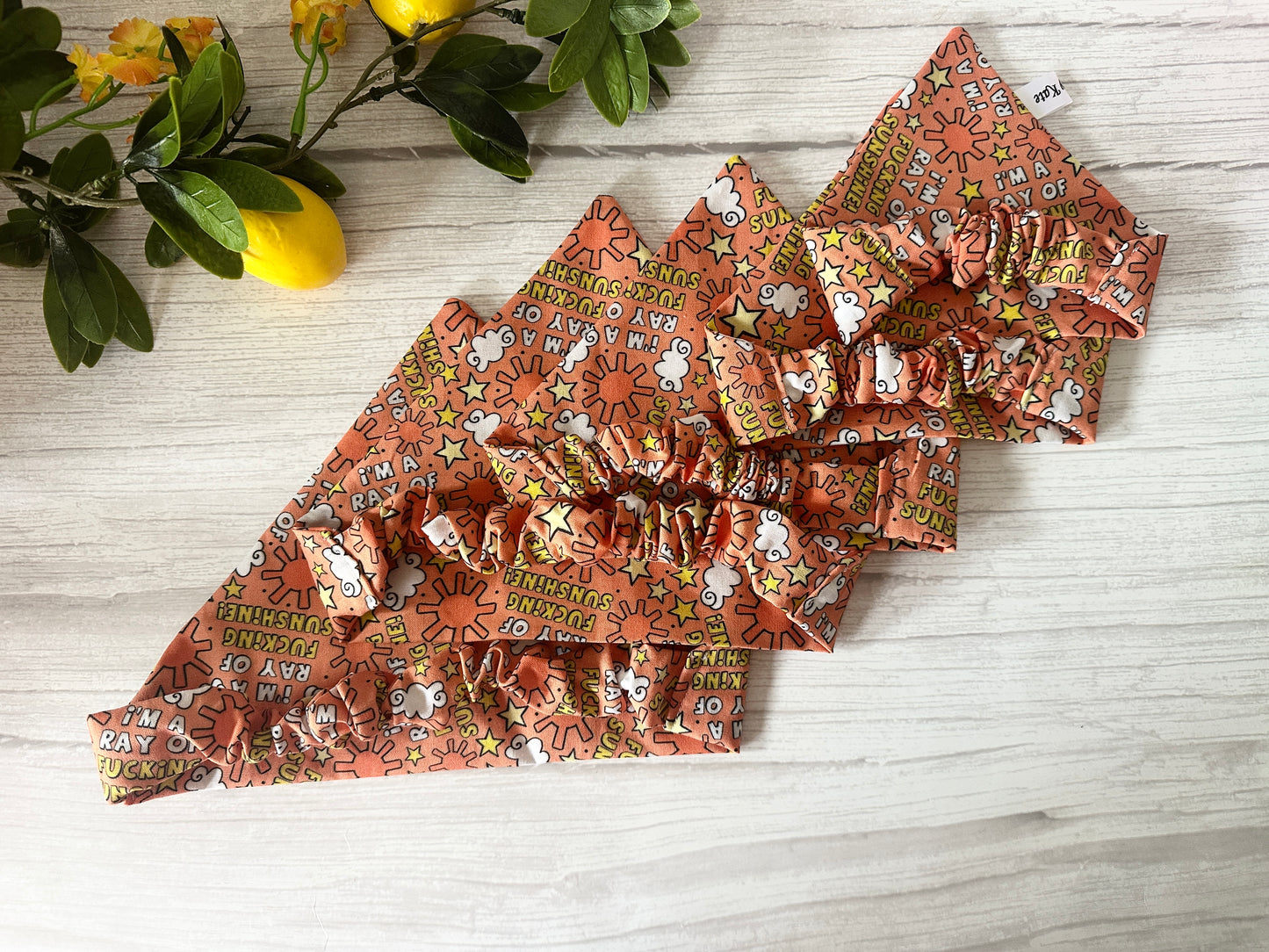 Four orange "I’m a ray of f***ing sunshine" Cotton Pet Scrunchie Bandanas by Crafts by Kate, with ruffled edges and sun/cloud prints, double as bandanas or headbands. Shown on light wood near green leaves and lemons.