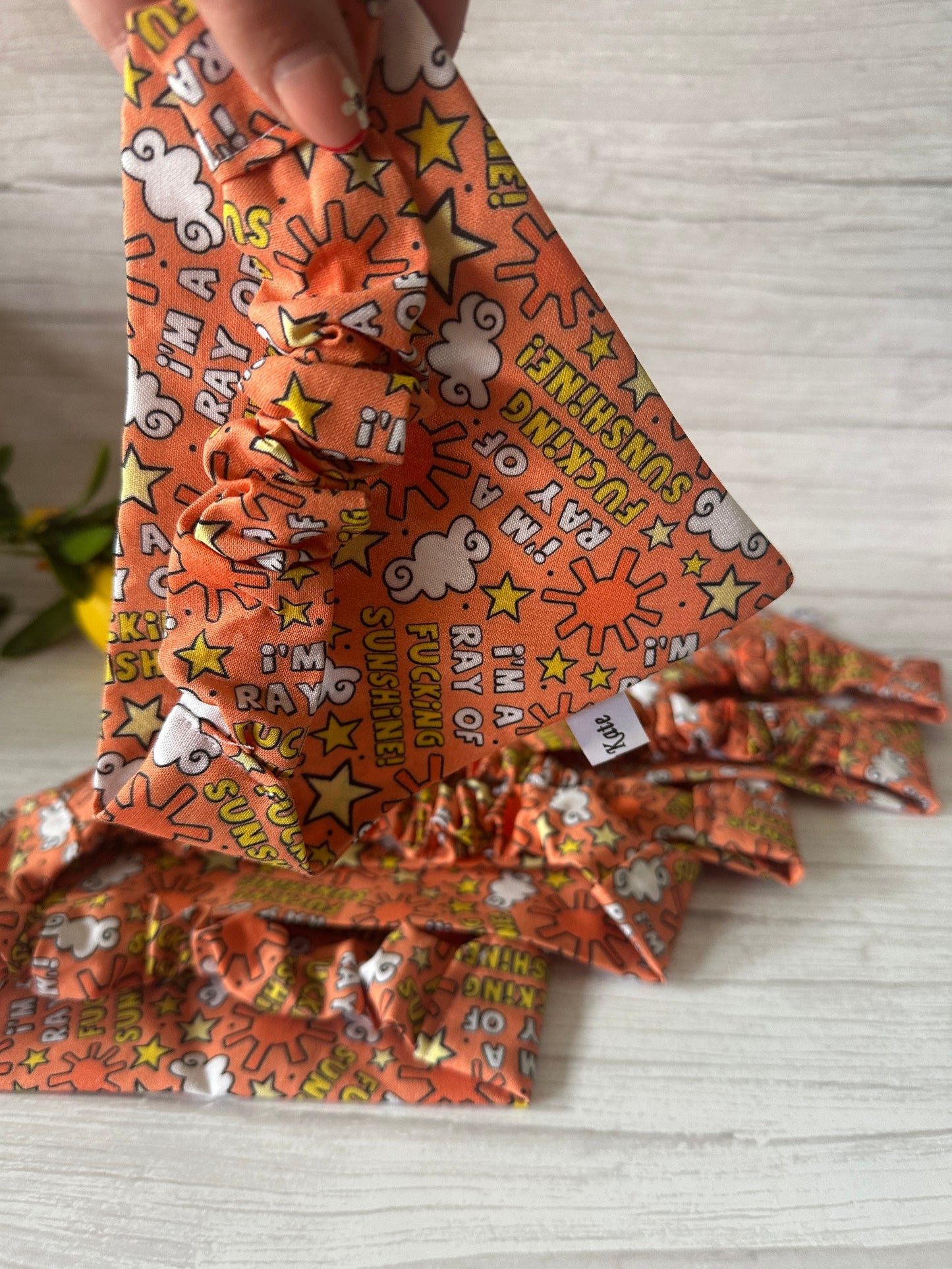 A hand holds the Crafts by Kate Cotton Pet Scrunchie Bandana – “I’m a ray of f***ing sunshine” – featuring cheerful sun, cloud, and star designs on a light wooden surface.