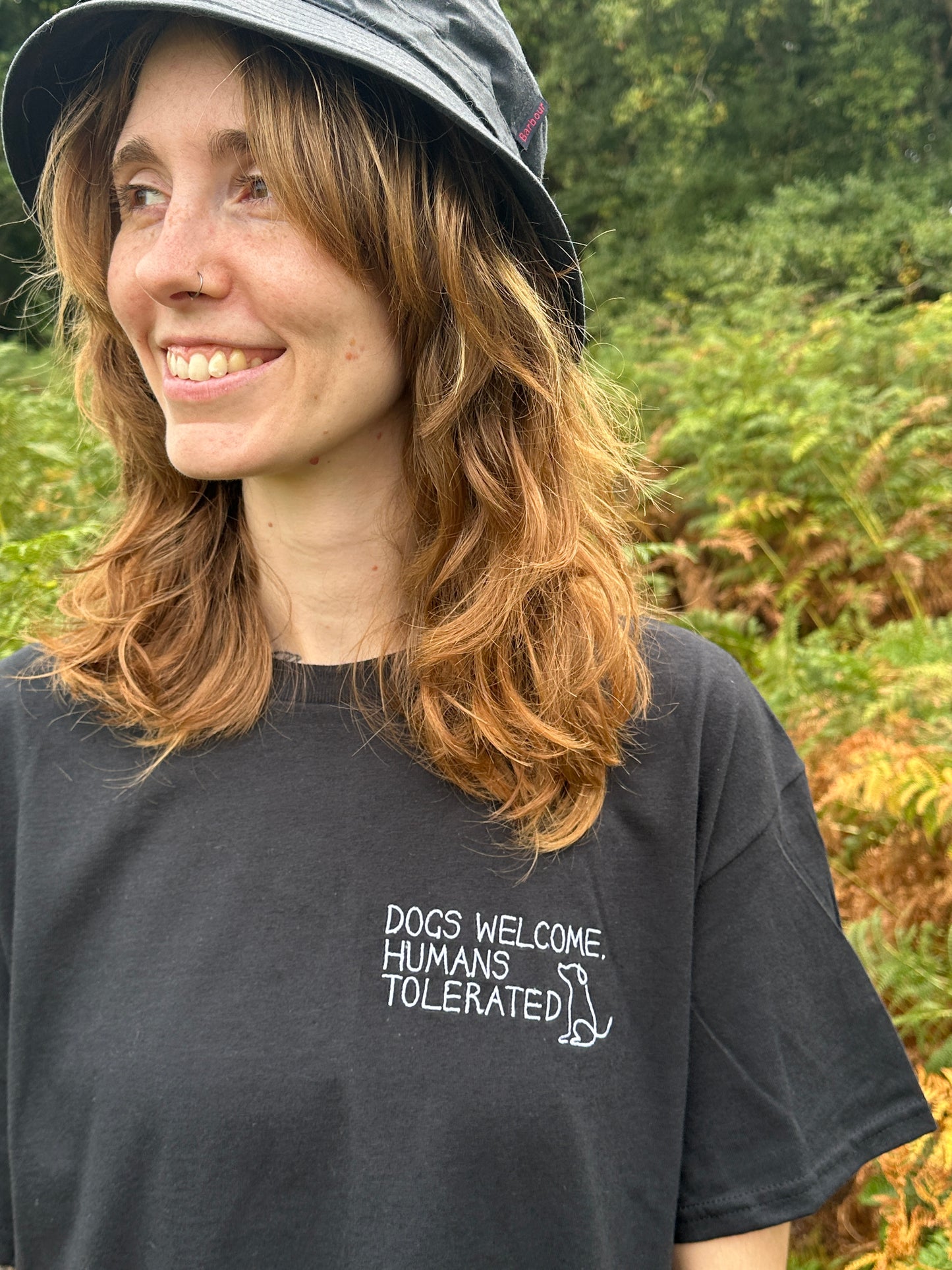 A smiling person with wavy hair wears the Crafts by Kate Dogs Welcome t-shirt—an oversized black unisex cotton tee with “DOGS WELCOME, HUMANS TOLERATED” and a small dog graphic—plus a black bucket hat, surrounded by lush green foliage.