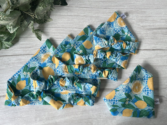 The Cotton Pet Scrunchie Bandana - Italian Lemons by Crafts by Kate features a matching blue and yellow lemon pattern, including bandanas, bow ties, and a handcrafted cotton pet scrunchie, shown on a light wooden surface with green leaves.