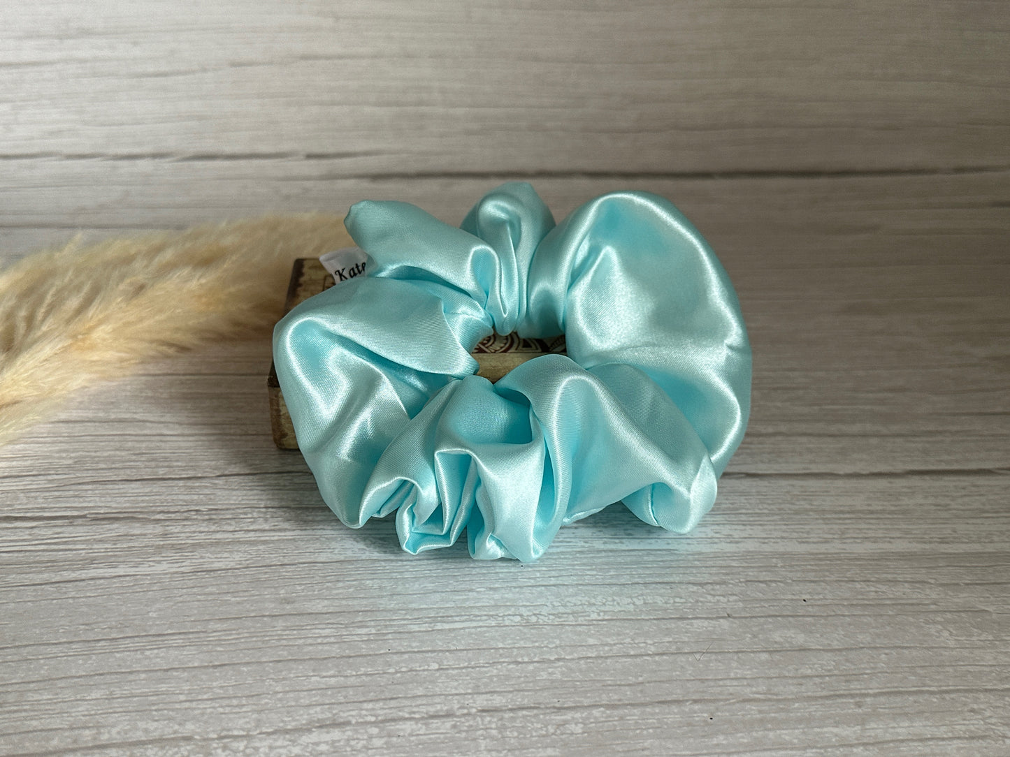 The Silk Extra Large Scrunchie - Aqua Blue from Crafts by Kate is showcased against a wooden backdrop, accompanied by a small arrangement of beige dried grass. The scrunchie's smooth and shiny silk material enhances its elegance as a sophisticated hair accessory.