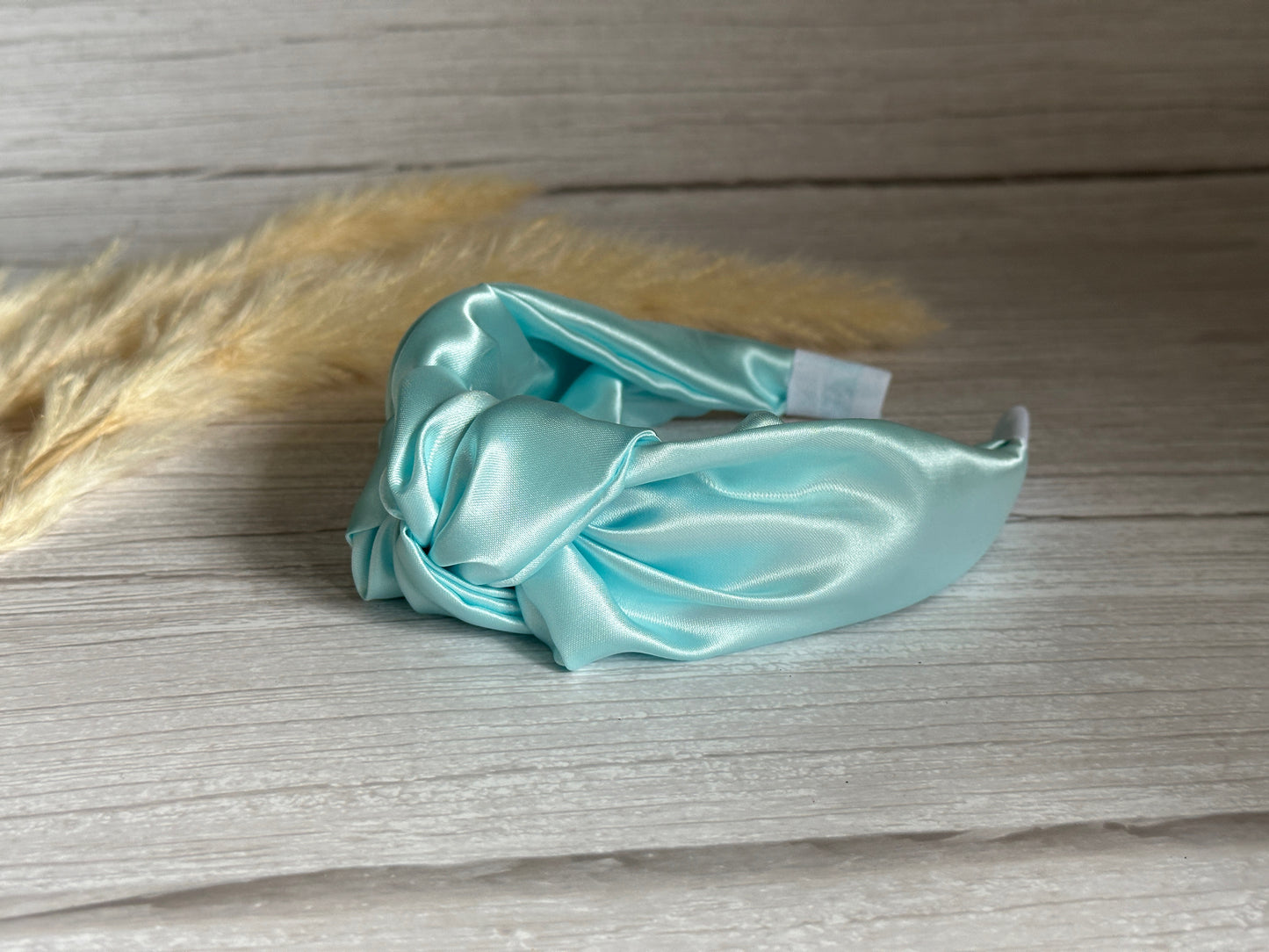 The Silk Knotted Headband - Aqua Blue by Crafts by Kate, a luxurious handmade piece in a light blue silk satin with a twisted knot design, rests elegantly on a wooden surface. Surrounding it are delicate wisps of beige pampas grass, adding to the sophisticated ambiance and making it an ideal accessory for enhancing any outfit with elegance and style.