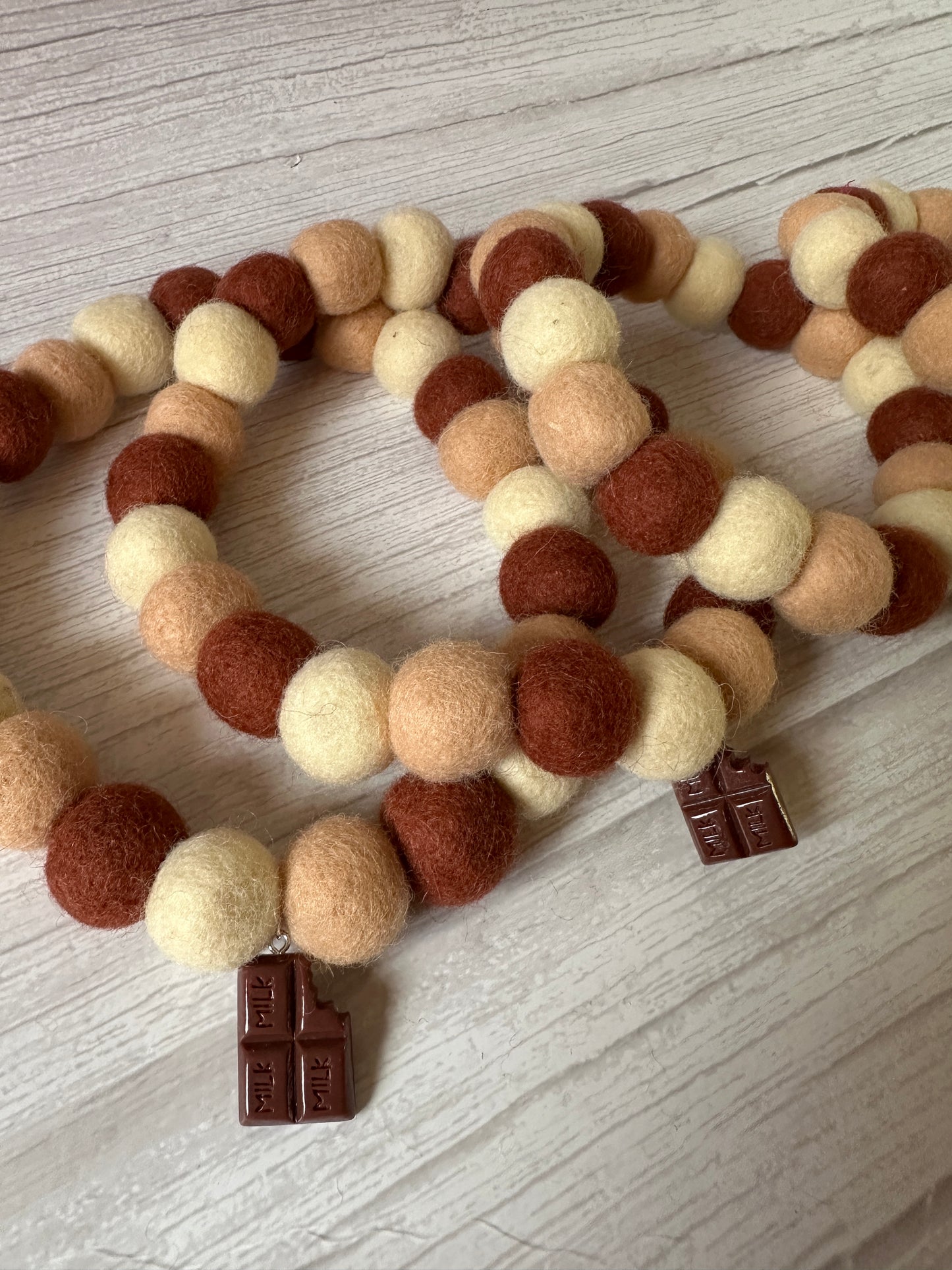 Three PomPom Pet Collars by Crafts by Kate, made of felt balls in cream, brown, and tan shades, are arranged on a wooden surface. Each collar echoes the Chocolate Lover design and features a small, rectangular tag.