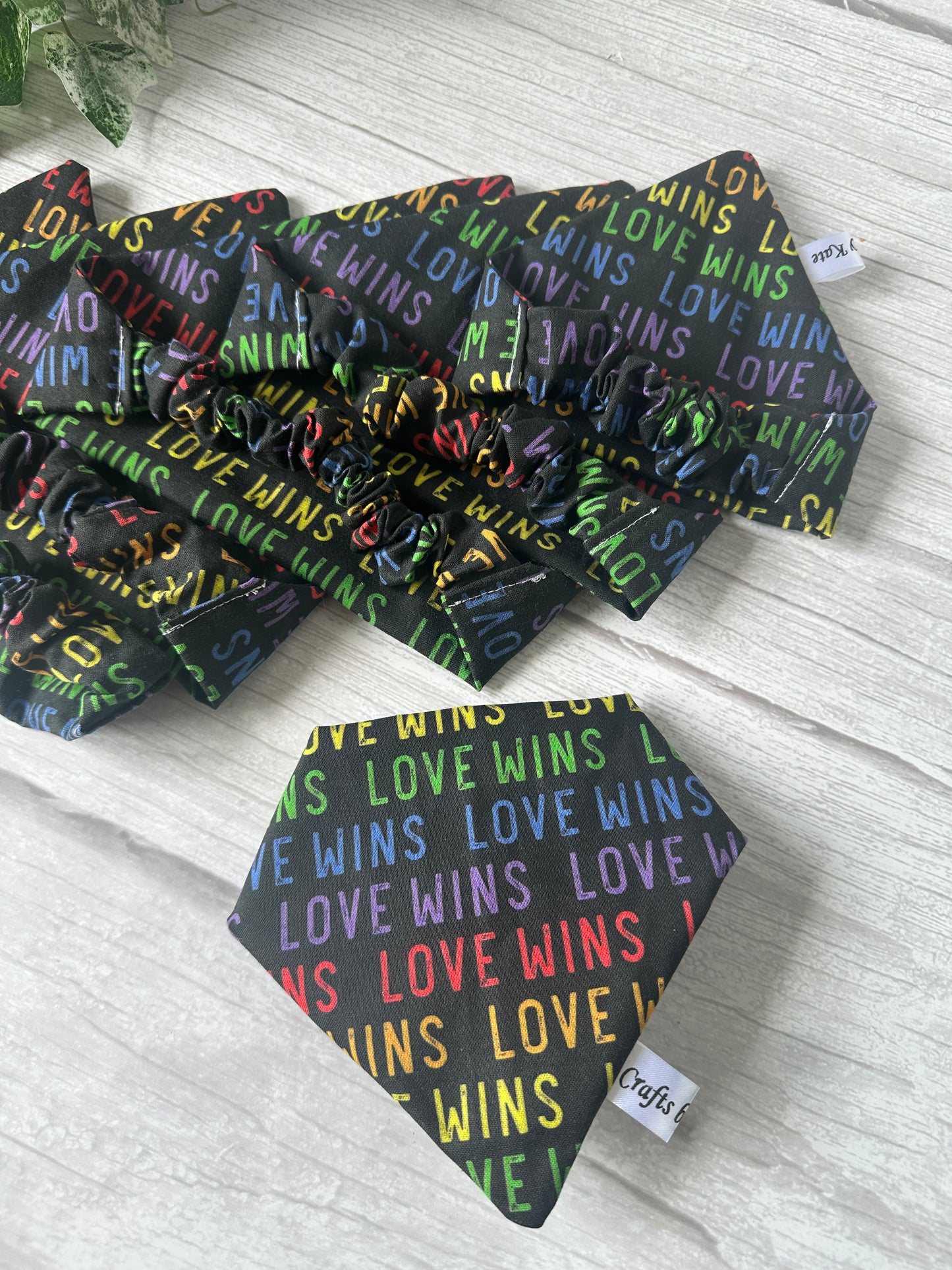 A selection of Crafts by Kate Cotton Pet Scrunchie Bandanas – Love Wins, featuring colorful "LOVE WINS" text celebrating the LGBTIQA+ community, are displayed on a light wooden surface with one bandana showing a white tag in front.