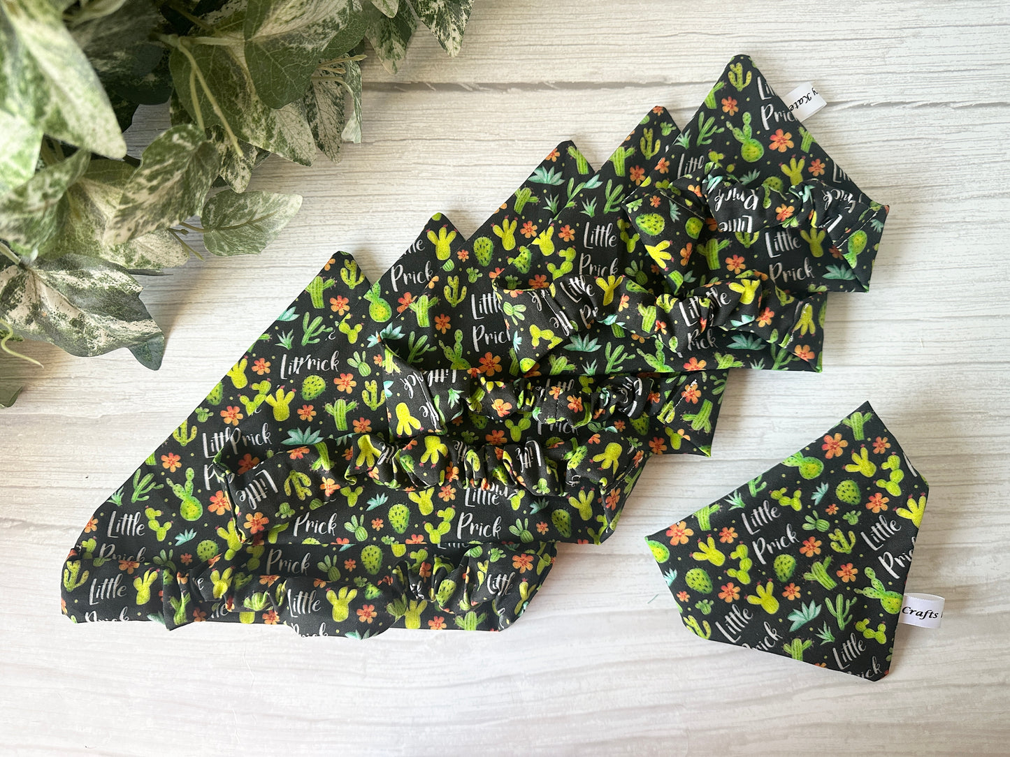 Five handcrafted black Cotton Pet Scrunchie Bandanas by Crafts by Kate, featuring colorful cactus and "Little Prick" patterns, are arranged on light wood with green plants—perfect accessories for stylish pets.