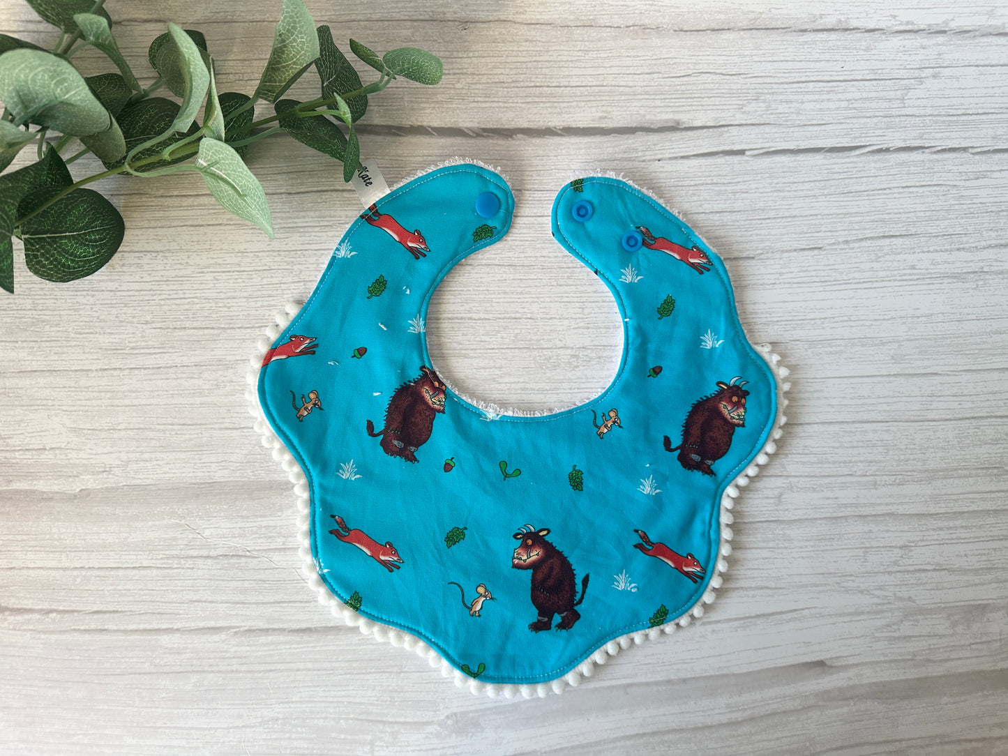 The Gruffalo in Blue baby gift set by Crafts by Kate includes a blue bib with mythical creatures, foxes, and leaves. Made from ultra-soft cotton with scalloped edges and dual snap buttons, it's ideal for a handcrafted collection. Features a small green plant in the upper left corner on light wood.