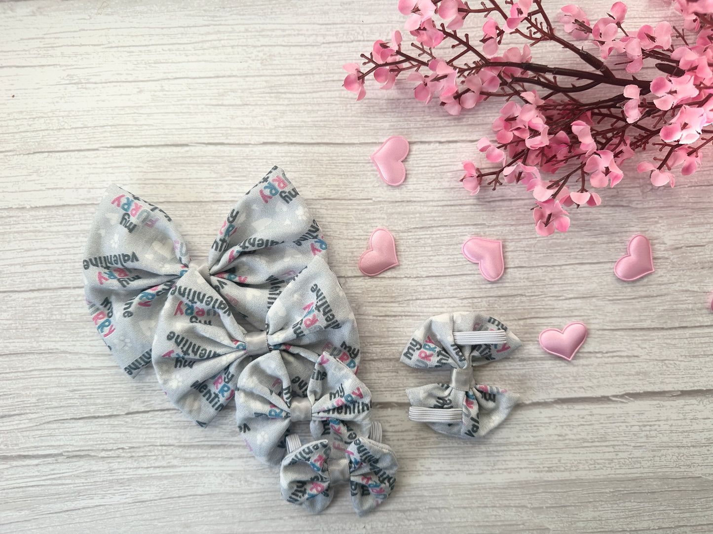 Crafts by Kate's "Cotton Pet Bow with collar attachment - My Fluffy Valentine" sits alongside pink artificial flowers and heart-shaped decorations on a light wooden surface.