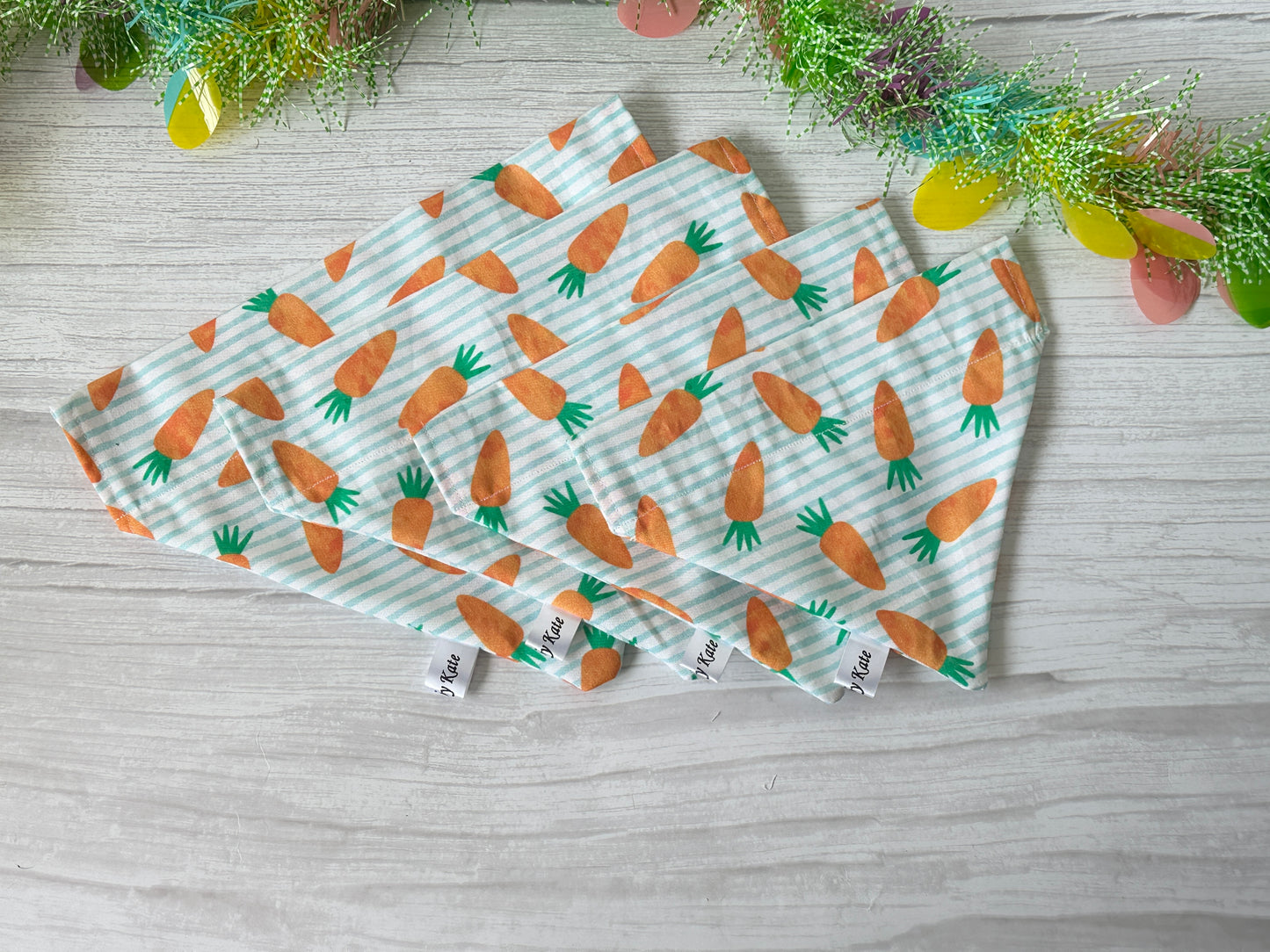 Four "Crafts by Kate" cotton pet bandanas featuring striped carrot designs rest on a light wooden surface, elegantly framed above by a handcrafted, colorful fringed garland with small pink, green, and yellow decorations.