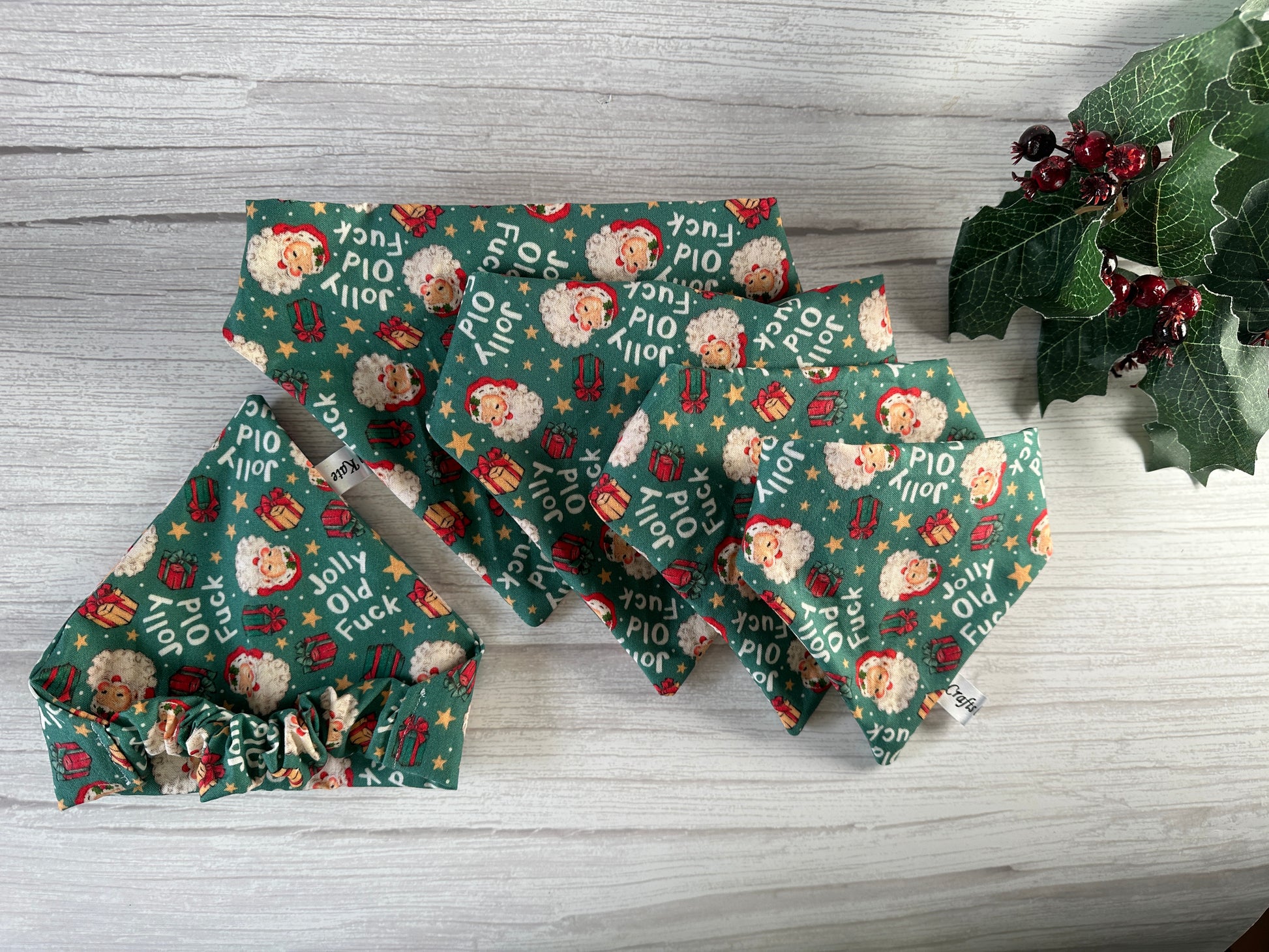 Assorted green fabric items with Christmas patterns, featuring Santa faces and words like "Jolly" alongside "Fk." A Cotton Pet Scrunchie Bandana - Jolly Old F*** by Crafts by Kate adds charm, displayed on a light wood surface with holly leaves and red berries.