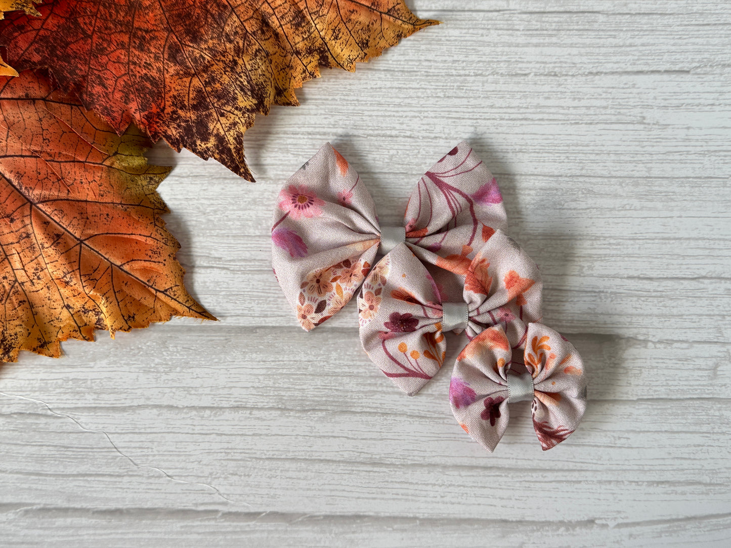 Three Crafts by Kate Cotton Pet Bows with collar attachment in woodland floral pink and orange designs are arranged on a light wooden surface with autumn leaves, highlighting their easy-to-wear elastic feature.