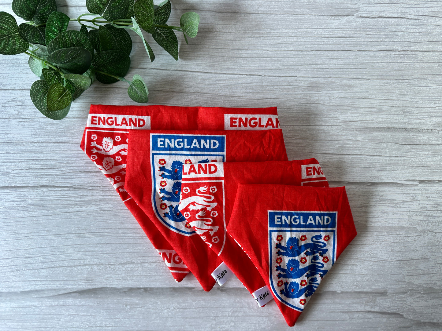 Three Crafts by Kate Cotton Pet Scrunchie Bandanas, each in different sizes and featuring an England football print with a lion crest, are displayed on a gray wooden surface. A small green plant in the upper left corner adds a natural touch.