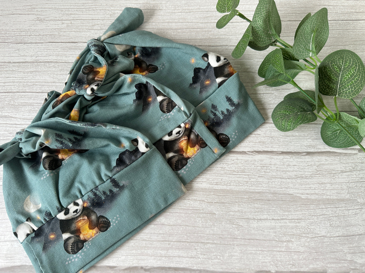 Two Baby Wool Cotton Jersey Knotted Hats in Panda Love design by Crafts by Kate are displayed flat on a light wood surface, with a leafy green plant to the right for an elegant touch.