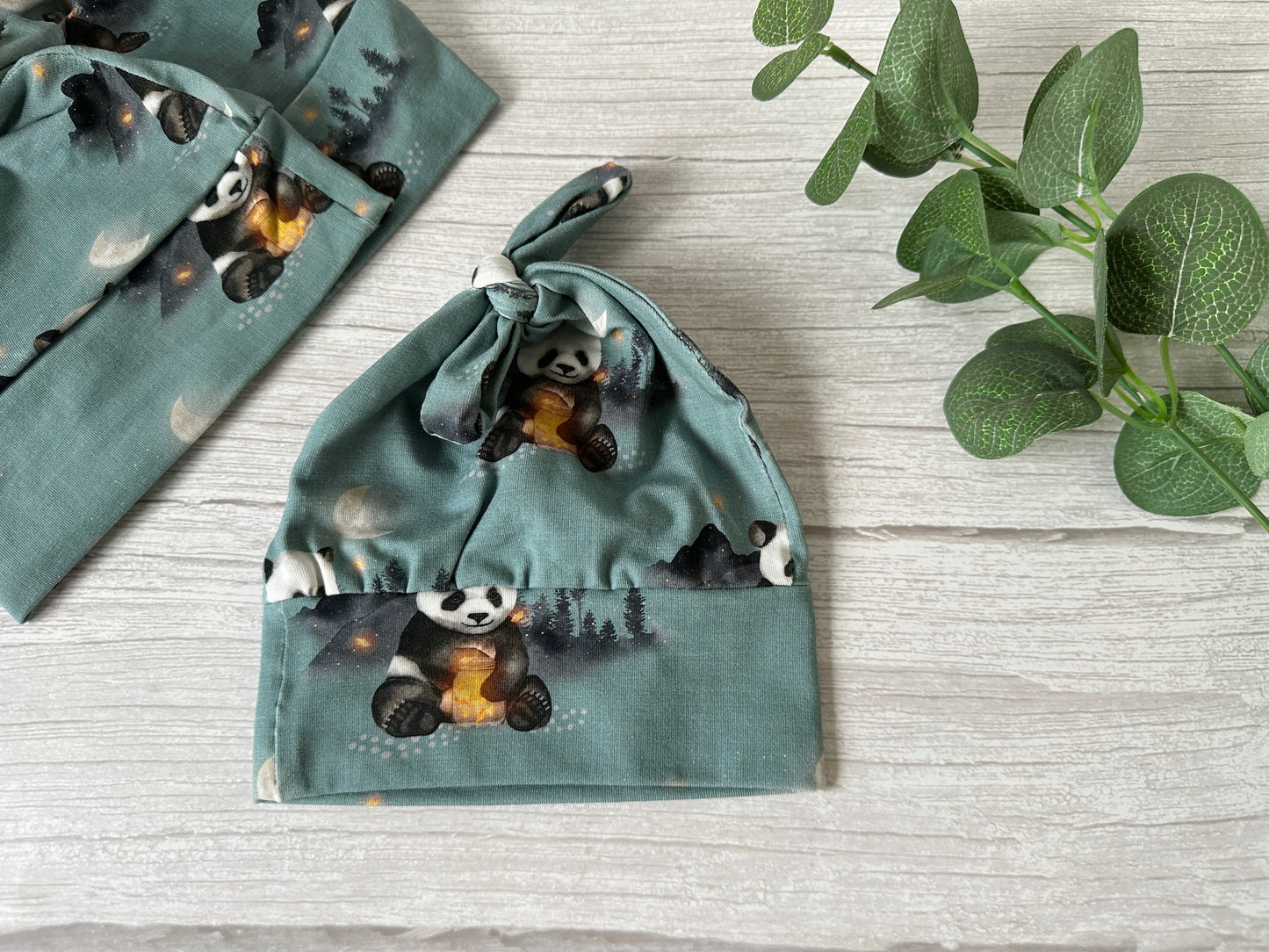 A Crafts by Kate Baby Wool Cotton Jersey Knotted Hat in green, adorned with pandas and trees, is displayed on a wooden surface with matching Panda Love Fabric; a small green plant adds a natural touch.