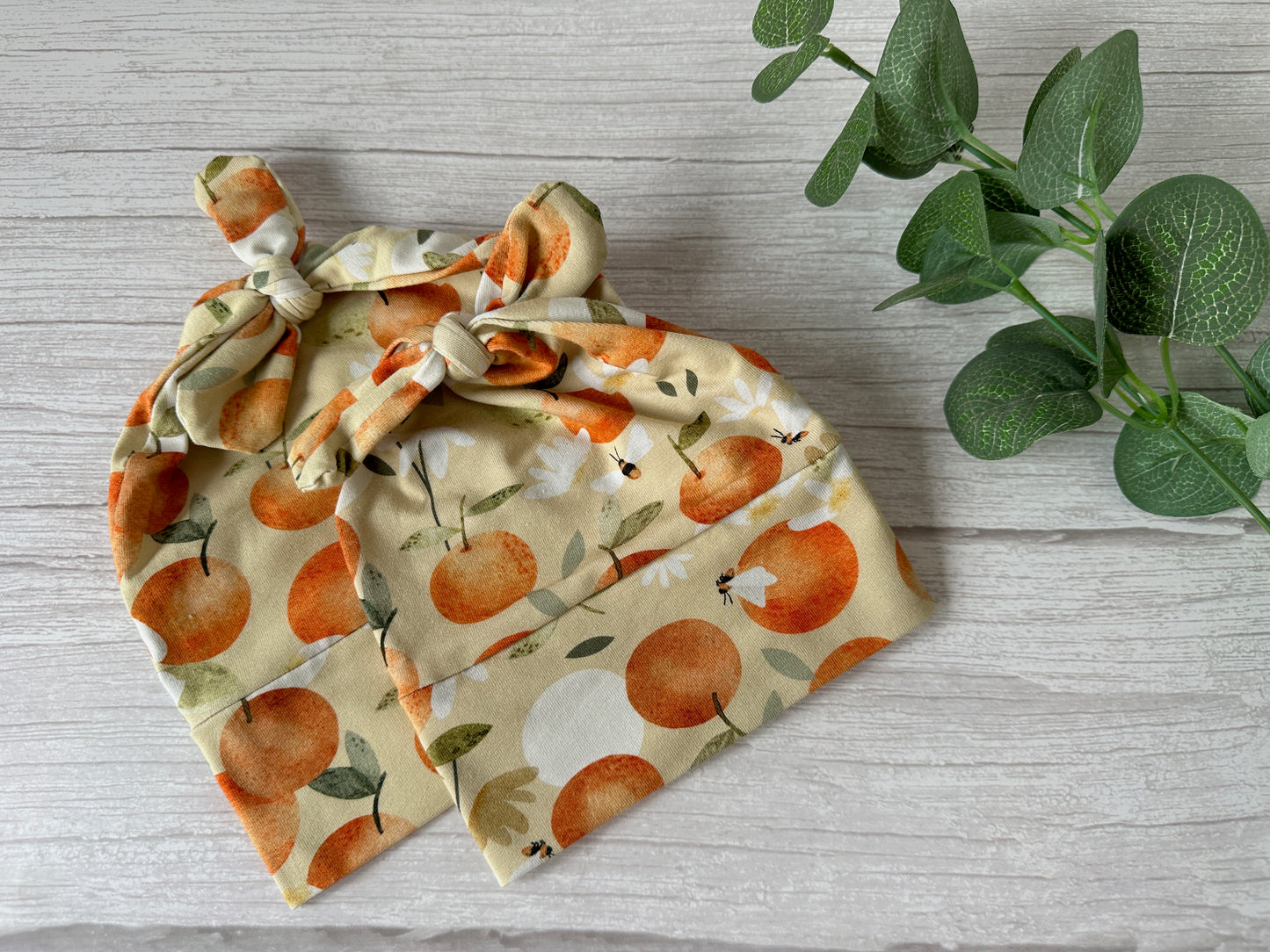 The "Baby Wool Cotton Jersey Knotted Hat - Summer Peaches" by Crafts by Kate features a tied knot design with orange fruits and leaves on a light background, beautifully presented on a light wooden surface alongside green leaf sprigs.