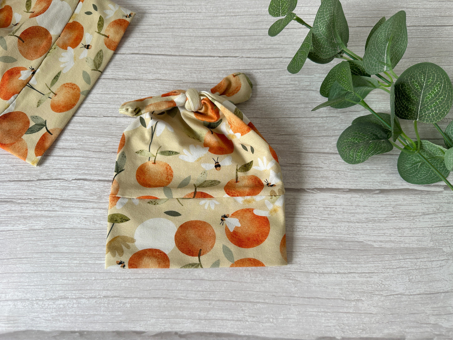 The "Summer Peaches" Baby Wool Cotton Jersey Knotted Hat by Crafts by Kate features handcrafted beige luxury wool with a tie knot, orange fruits, brown bees, and white daisies. It's displayed on a light wooden surface with green leaves and matching fabric.