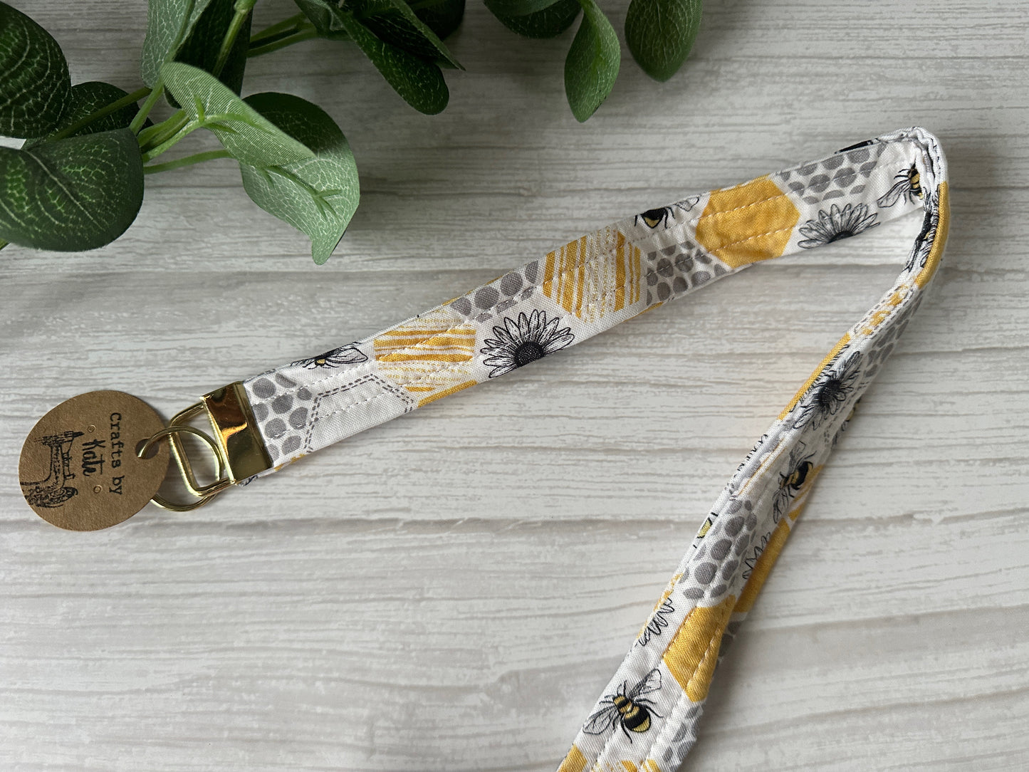 The Cotton Lanyard with Metal Fob Keychain - Beehive by Crafts by Kate rests on a light wooden surface, showcasing yellow, gray, and white floral patterns with bees. A visible tag reads "crafted by." Green leaves peek from the corner.