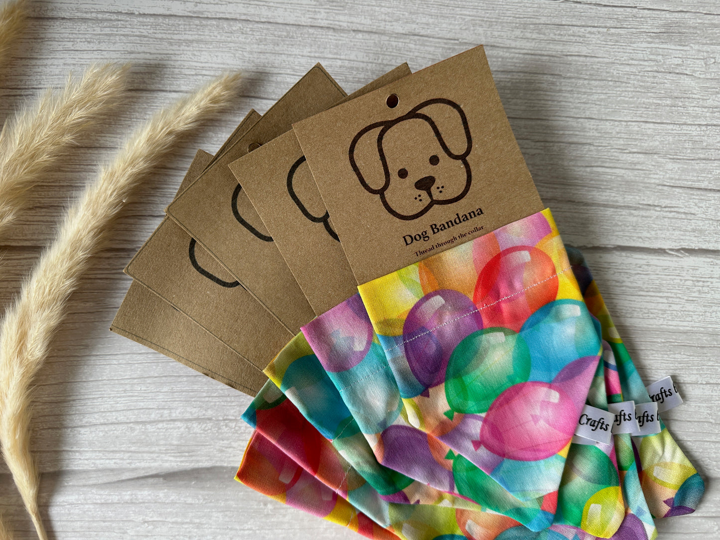 The "Birthday Balloons" Cotton Pet Bandanas by Crafts by Kate are neatly arranged, each handcrafted from cotton with a vibrant balloon pattern. Each bandana comes with a brown cardboard tag showcasing an illustration of a dog's face and the text "Dog Bandana." Dried wheat stalks are placed beside these pet accessories on a light wooden surface.