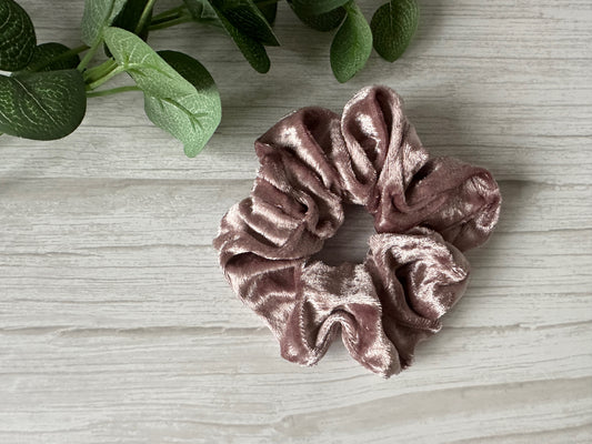 The Crushed Velvet Extra Large Scrunchie in Blush Pink by Crafts by Kate rests on a light wooden surface beside green leafy stems, showcasing its handcrafted luxury design.