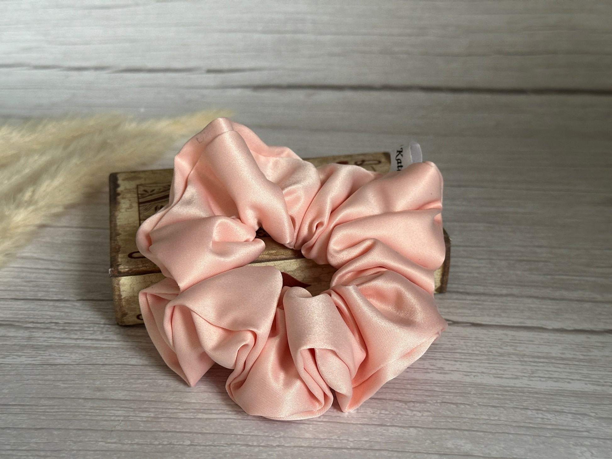 A pink satin scrunchie from Crafts by Kate's Satin Extra Large Scrunchies - Bridal Collection rests on a small wooden box on a light wooden surface. Two light-colored feathers softly appear in the background, enhancing the arrangement's delicate touch.
