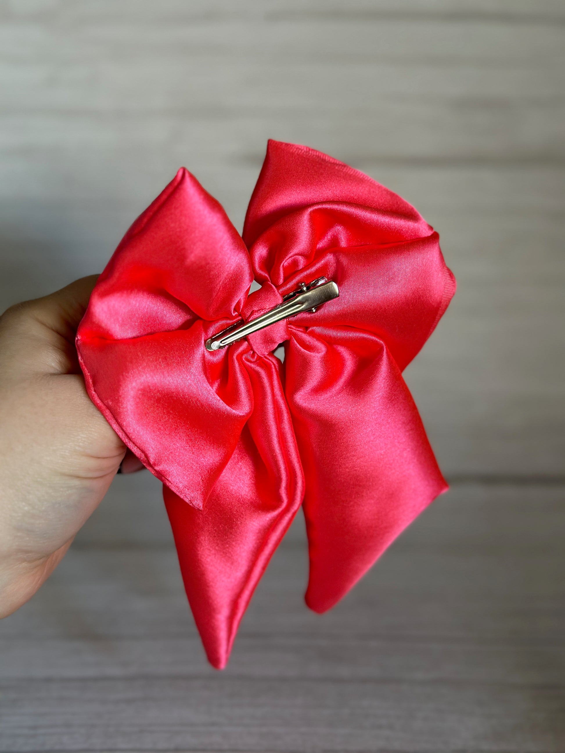 A hand cradles a luxurious Silk Sailor Bow Clip from Crafts by Kate, its vibrant bright pink silk glistening against a blurred wooden backdrop. This handcrafted accessory features a sleek metal clip, perfect for adding an elegant touch to any hairstyle.