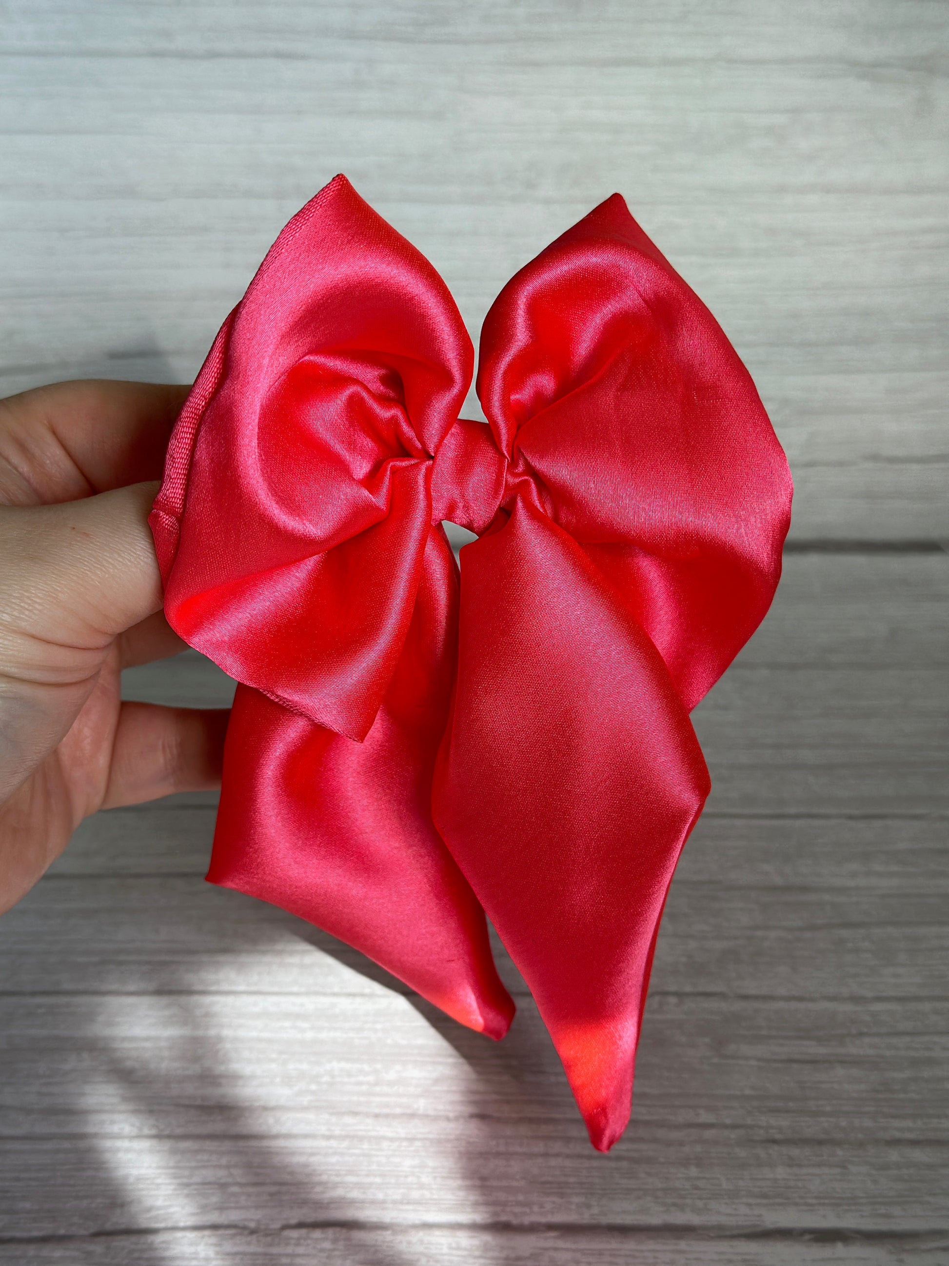 A hand holds a luxurious sailor bow, its shiny bright pink silk contrasting beautifully against a wooden background, showcasing the vibrant color and smooth texture of this handcrafted accessory by Crafts by Kate.