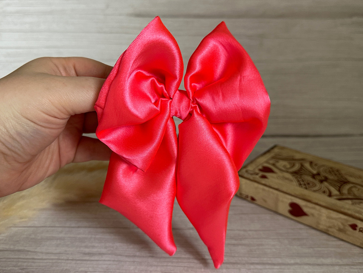 A hand holding a luxurious Silk Sailor Bow Clip in vibrant bright pink satin against a wooden background. A decorative wooden box with heart designs is partially visible next to this exquisite, handcrafted accessory from Crafts by Kate.
