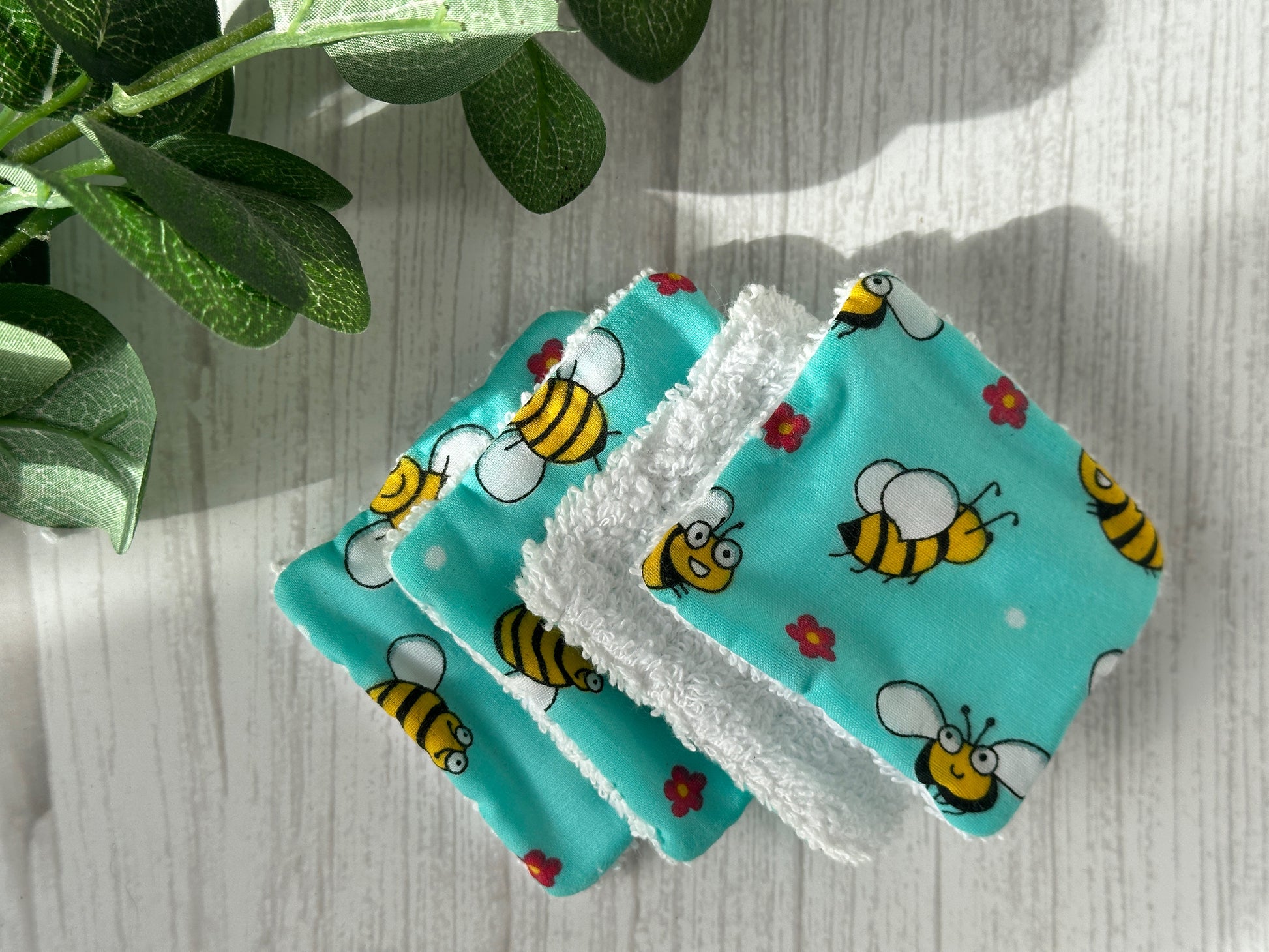 A set of Crafts by Kate's Cotton Reusable Make-up Remover Pads - Bumble Bee is arranged on a light wooden surface. These environmentally friendly, handcrafted cloth wipes feature clean illustrations of cheerful bees and small red flowers on a turquoise background. Fresh green leaves partially frame the image from the upper left corner.
