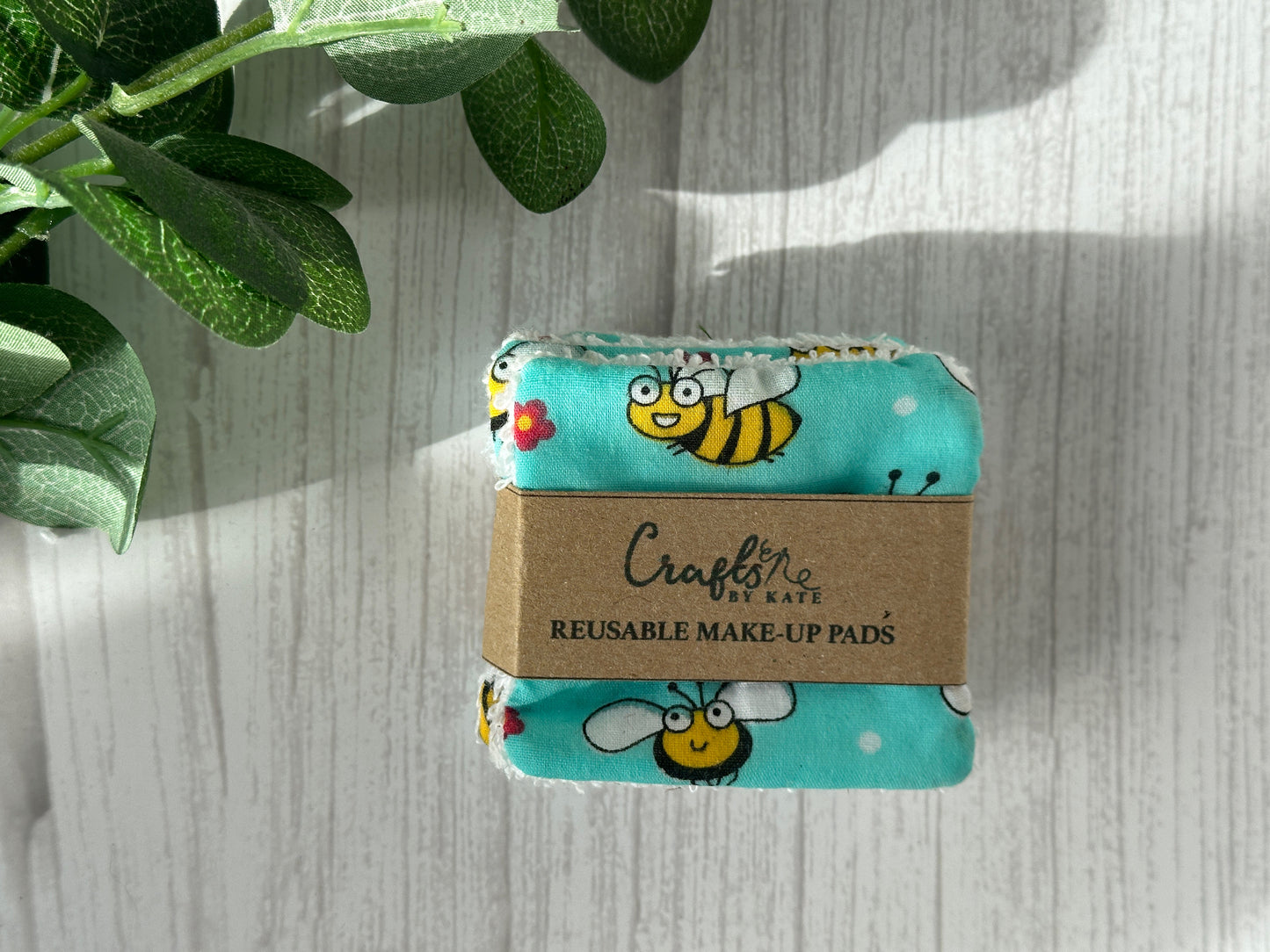 A set of "Cotton Reusable Make-up Remover Pads - Bumble Bee" by Crafts by Kate, crafted from cotton and terry, wrapped in a brown paper band with "Crafts by Kate" imprinted on it. The pads boast a vibrant blue fabric adorned with an adorable bee pattern. Green leaves are partially visible in the top left corner of the image, emphasizing its eco-friendly nature.