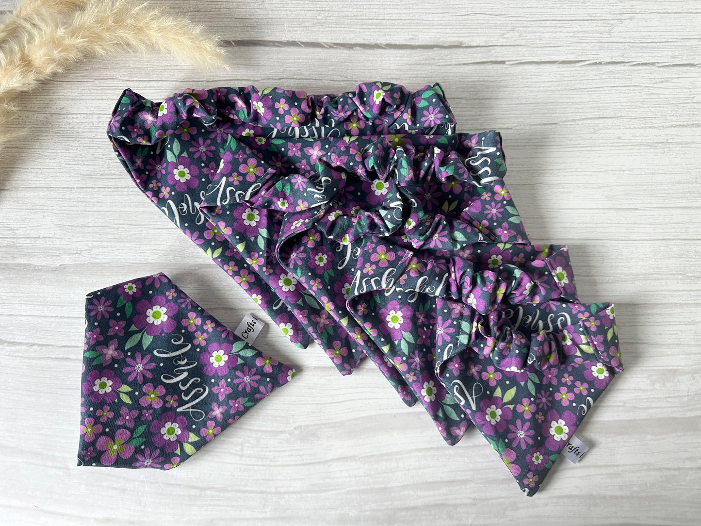 Four floral scrunchies in purple and green from Crafts by Kate, along with a matching Cotton Pet Scrunchie Bandana - Asshole angled to the bottom left with a visible tag, are elegantly arranged on a light wooden surface.