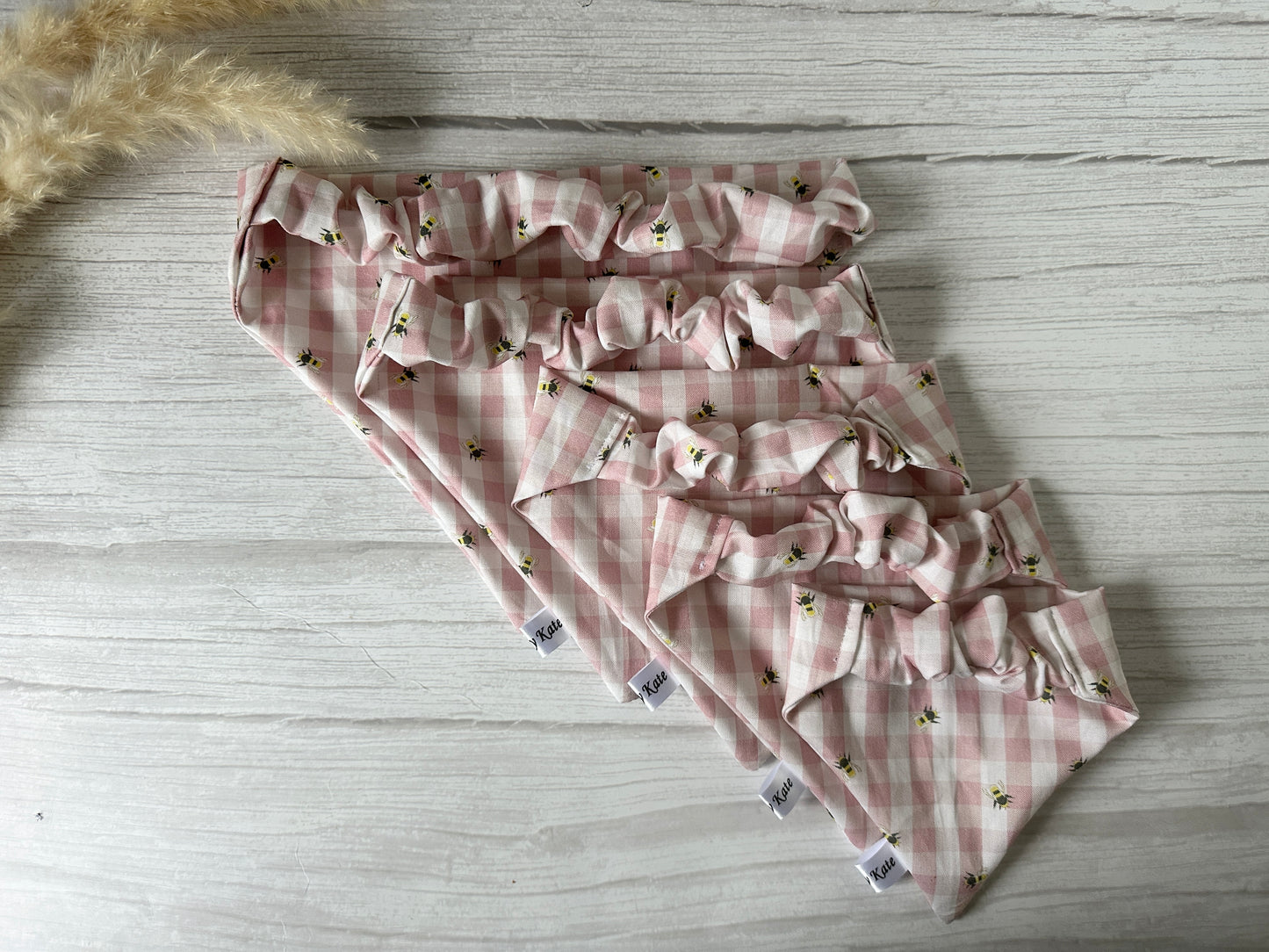 Four Cotton Pet Scrunchie Bandanas by Crafts by Kate, featuring pink gingham bees and small floral patterns, are neatly arranged on a light wooden surface with beige pampas grass in the top left corner.