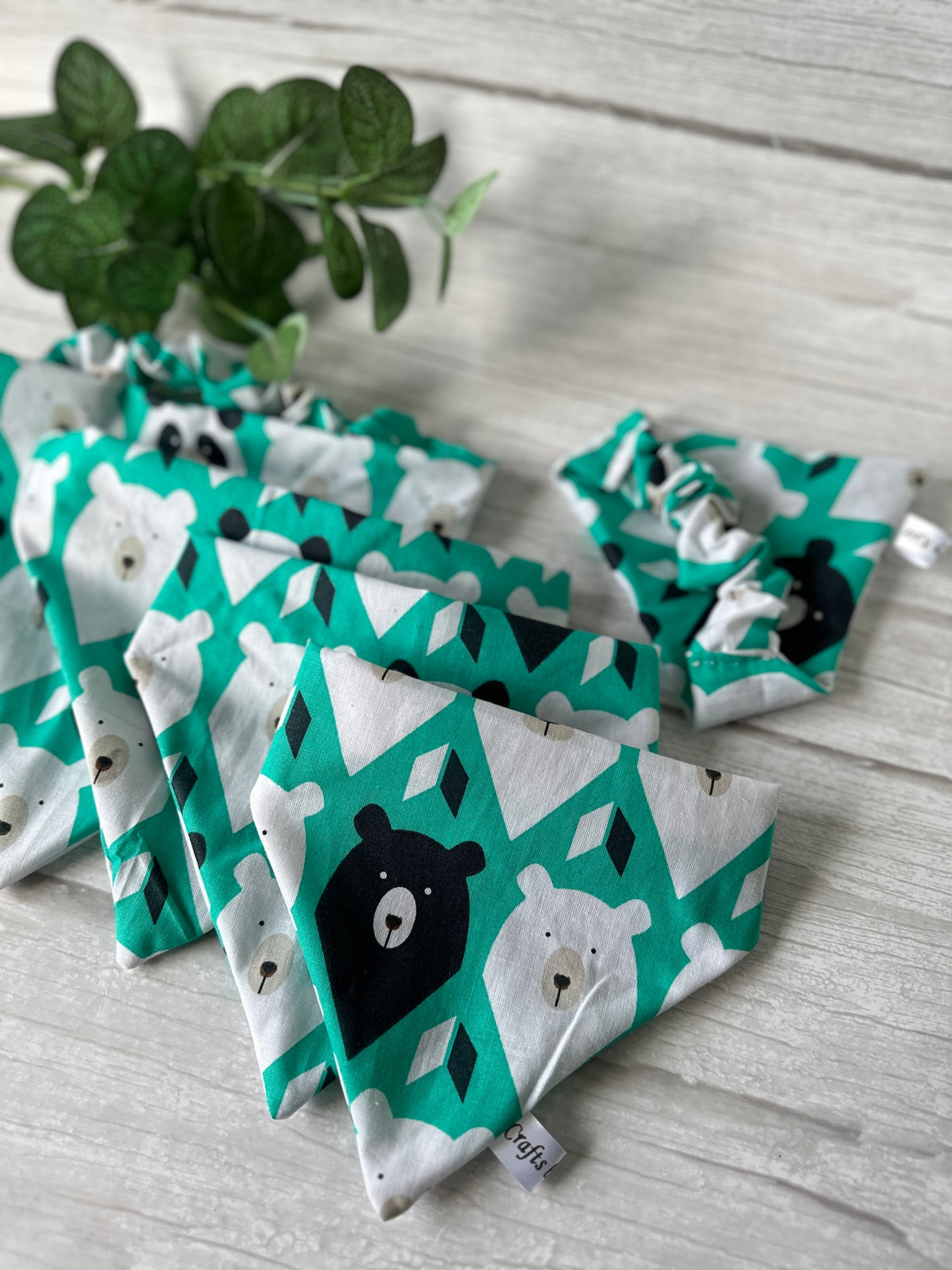 Five "Cotton Pet Scrunchie Bandanas - It’s My Barkday" by Crafts by Kate are neatly arranged on a light wooden surface. Each bandana showcases bear faces on a diamond-patterned green background, perfect for bear enthusiasts, with a small green plant in the top left corner to complete the look.