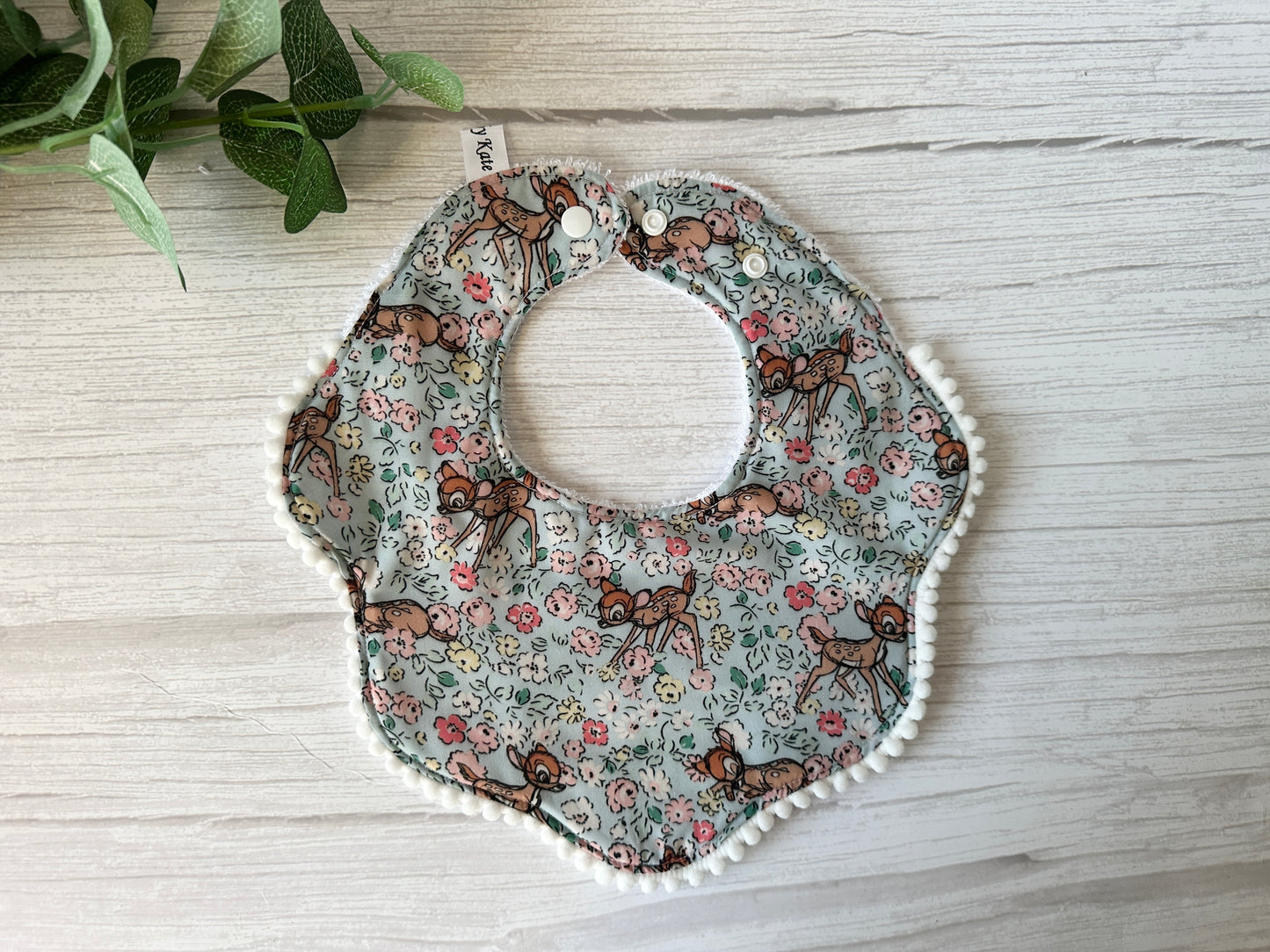 The Crafts by Kate Cotton & Terry Towelling Baby Gift Set - Floral Bambi features a bib with small deer, flowers, and leaves printed on ultra-soft cotton. It has two white snaps at the top. A plant with green leaves appears at the corner of a white wooden surface.