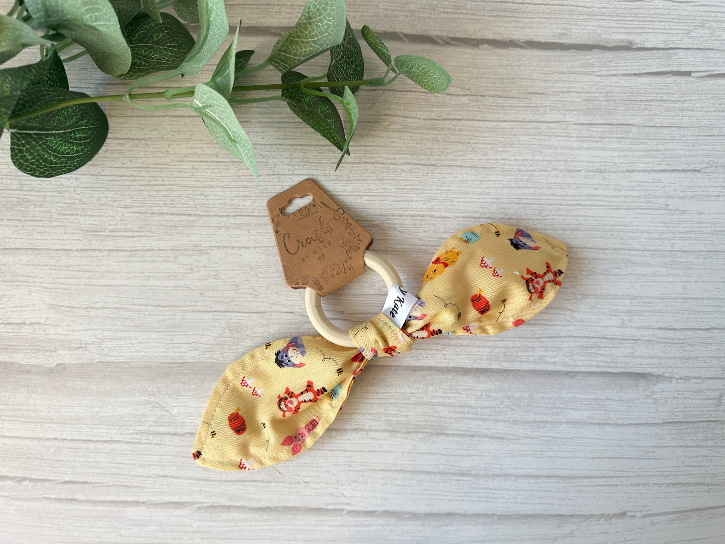 A charming baby gift set by Crafts by Kate, featuring a wooden teething ring with ultra-soft cotton in bow form, showcases colorful prints of animals and objects. Comes with a brown cursive tag on a white wooden backdrop with green leaves. Ideal for fans of the Winnie the Pooh & Friends collection!.