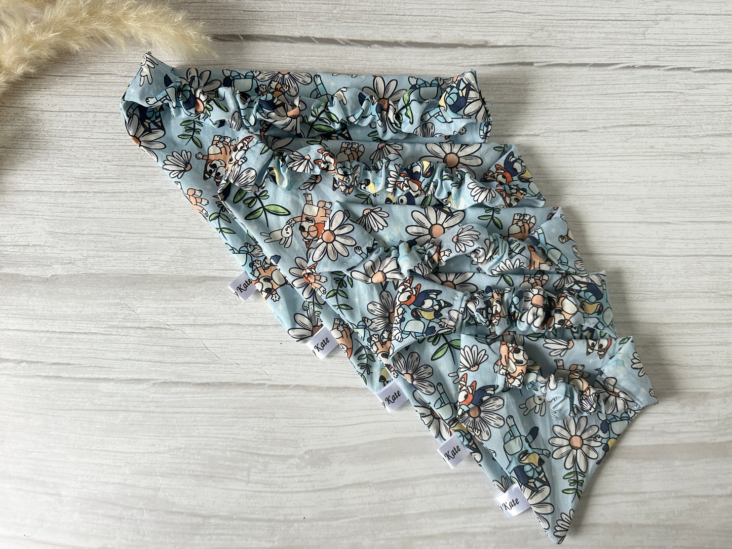 Five handcrafted Cotton Pet Scrunchie Bandanas in pastel blue, with ruffle details, from Crafts by Kate are fanned out on a white wooden surface. Each Bluey & Bingo piece features a Spring Tartan pattern of daisies and blooms, highlighting a delicate garden theme for your furry friend.