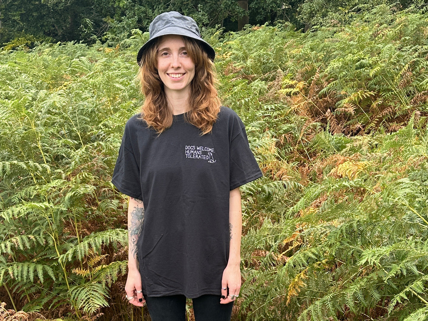 A smiling woman with wavy brown hair wears the Crafts by Kate "Dogs welcome" t-shirt in black and a bucket hat, standing outdoors in front of lush green ferns.