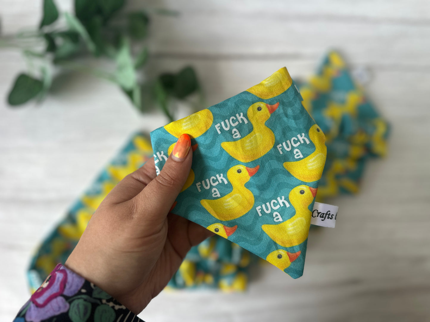 A hand displays the Crafts by Kate Cotton Pet Scrunchie Bandana, featuring a blue duck print and the phrase "F*** a duck." A leafy plant and matching fabric appear blurred in the background.