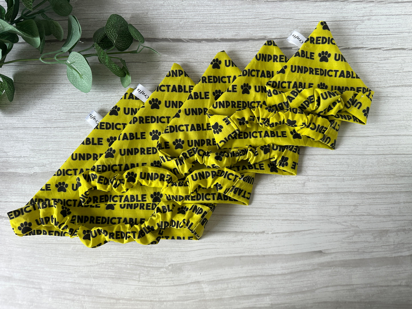 The Cotton Pet Scrunchie Bandana - Unpredictable by Crafts by Kate is a yellow, handcrafted accessory with black paw prints and “UNPREDICTABLE” printed repeatedly, shown on light wood with green leaves in the top left corner.