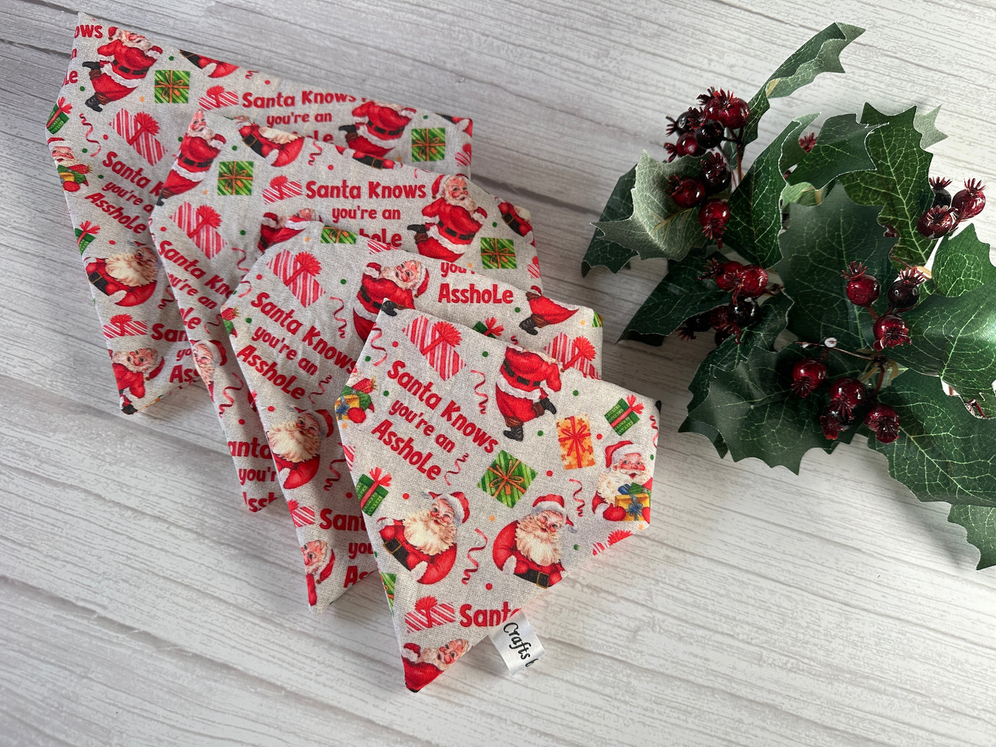The Cotton Pet Scrunchie Bandana - Santa Knows by Crafts by Kate features cartoon Santas, presents, and the cheeky phrase “Santa Knows you’re an Asshole,” with holly, displayed on a white wooden surface.