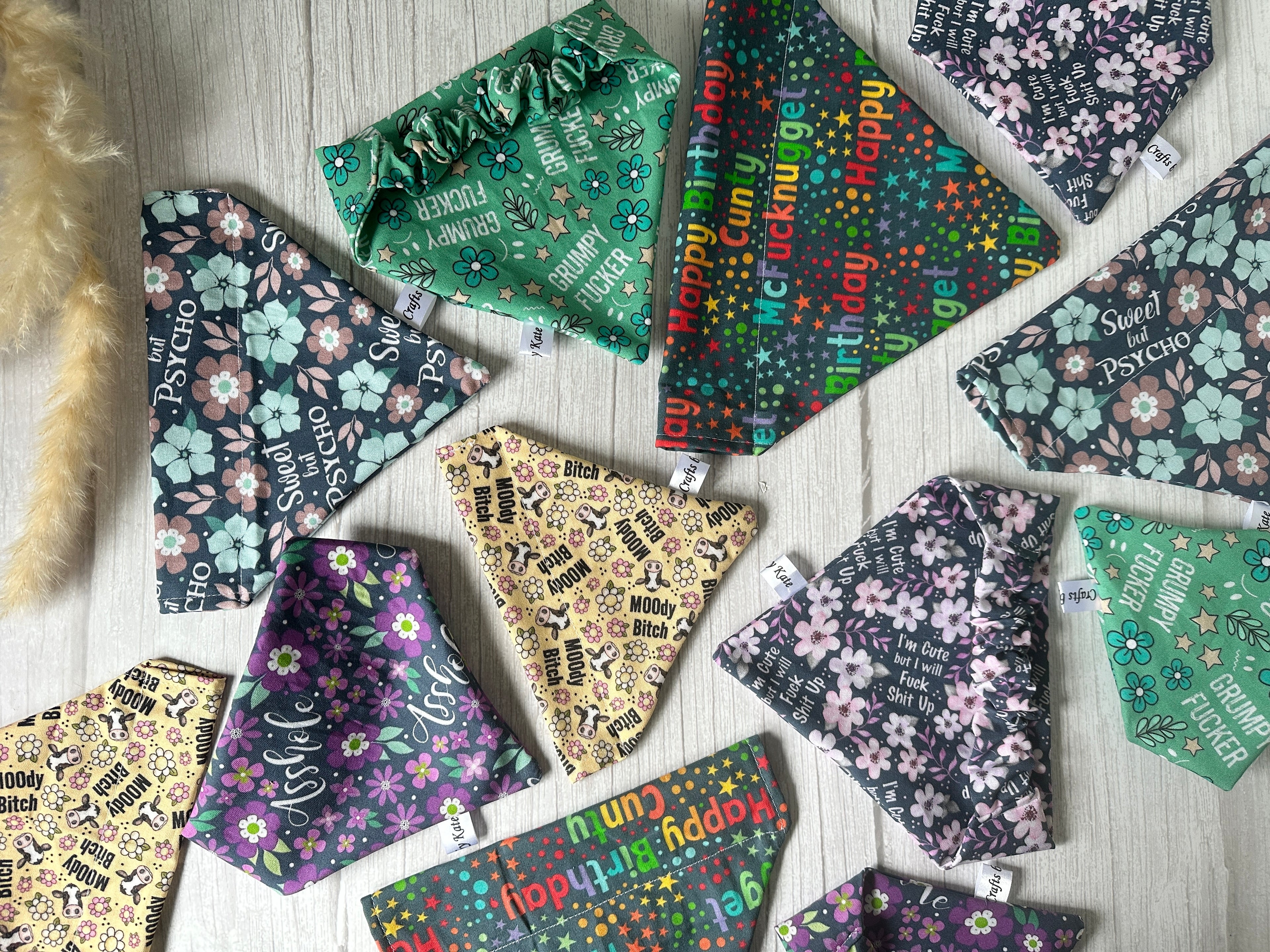 Colorful fabric pieces, featuring designs with phrases like "Happy," "Sweet," and "Moody Bitch," are spread across a white wooden surface. A fuzzy object wearing a handcrafted Crafts by Kate Cotton Pet Scrunchie Bandana - “I'm Cute but I will Fuck Shit up!” is partially visible on the left edge.
