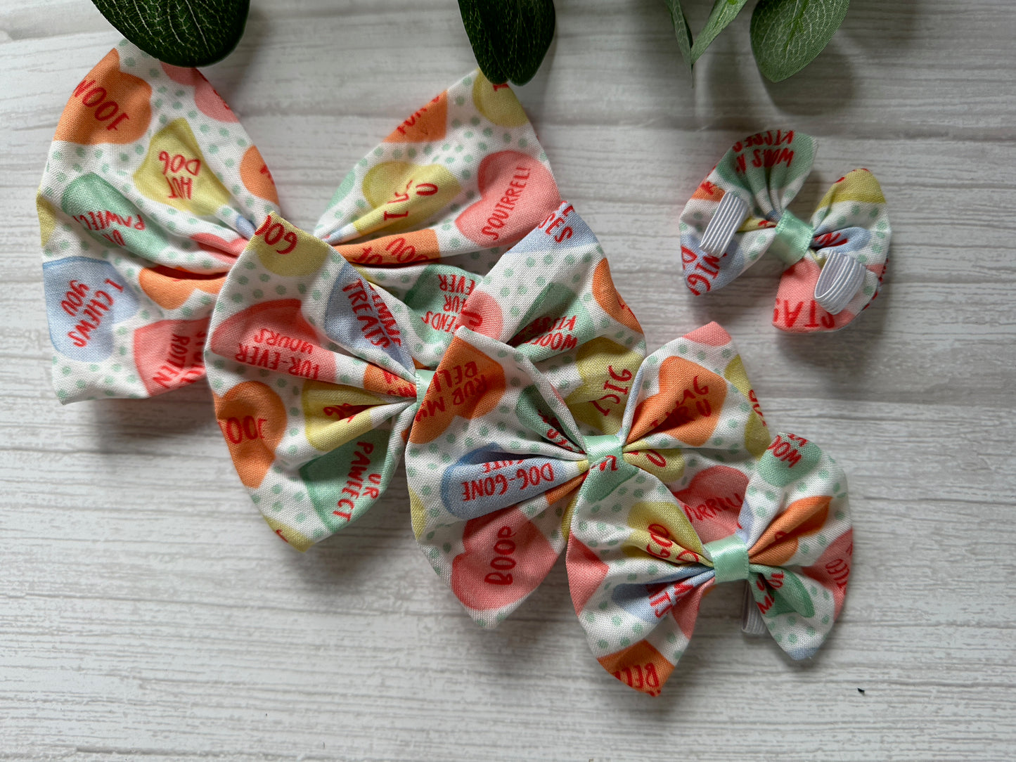 Crafts by Kate's Cotton Pet Bow with collar attachment showcases pastel heart patterns and phrases like "Top Dog," "Bone Appetite," and "Dig It." These charming accessories, featuring elastic fastening for convenience, are displayed on a light wooden surface adorned with green leaves.