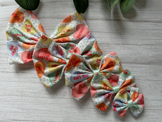 A vibrant, multi-layered bow from Crafts by Kate, crafted with Candy Love Hearts fabric in pastel shades. Featuring "BOO!" and "YOU'RE FAB" on a dotted backdrop, this Cotton Pet Bow with collar attachment rests elegantly on a light wooden surface accented by green leaves above.