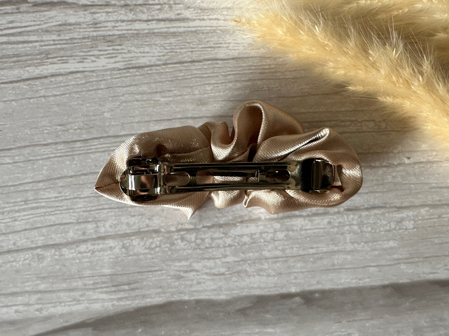 A close-up of the Silk French Barrette Scrunchie Clip - Champagne Gold by Crafts by Kate shows it attached to a luxurious silk scrunchie on a textured gray surface, with a fluffy light-colored object partially visible in the corner.