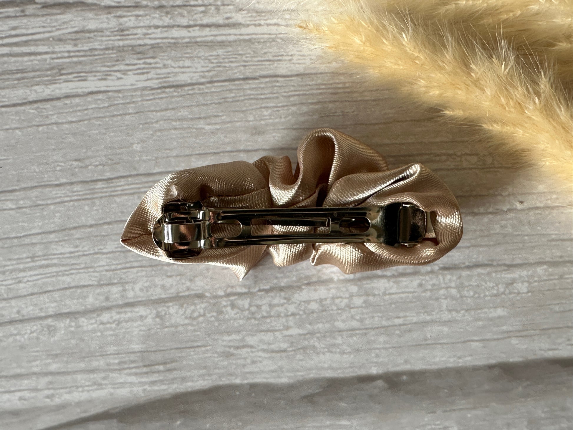 A close-up of the Silk French Barrette Scrunchie Clip - Champagne Gold by Crafts by Kate shows it attached to a luxurious silk scrunchie on a textured gray surface, with a fluffy light-colored object partially visible in the corner.